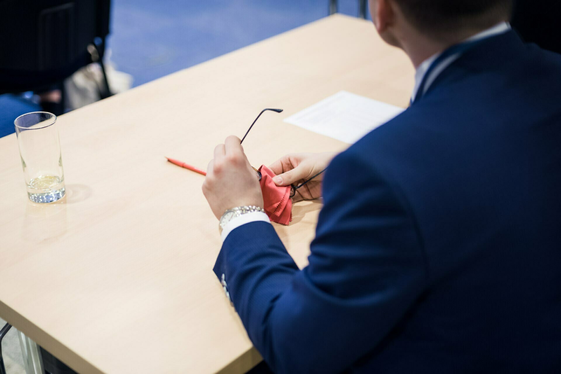 A person in a blue suit sits at a desk, holding glasses and a cleaning cloth, illustrating the finer details often captured in event photography. On the table next to him lies a pencil and an empty glass. The person's face is not visible, which adds to the intrigue of event photography.  