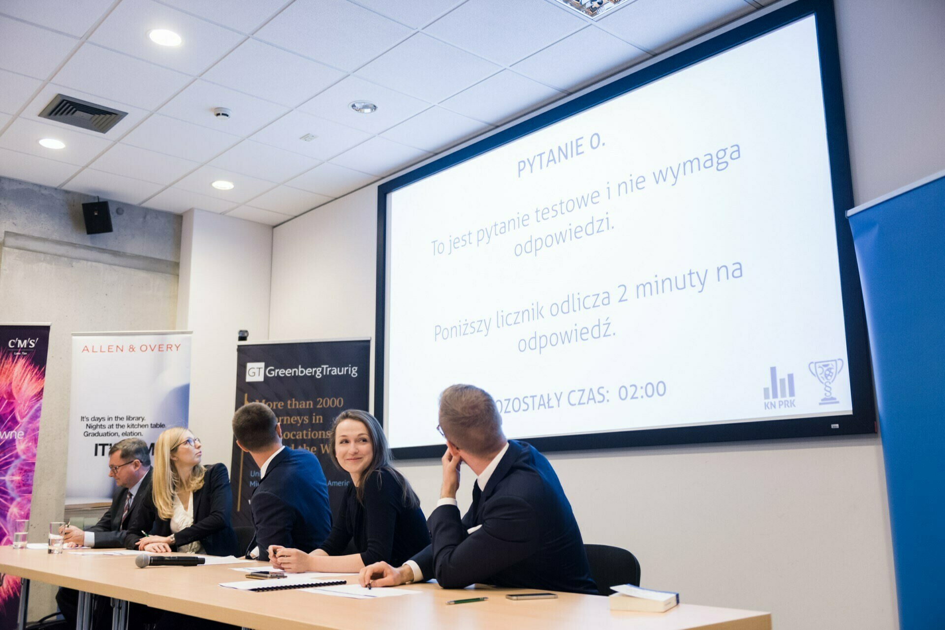 Five people, four men and one woman, sit at a table in a conference room. In the background is a projection screen displaying a slide with Polish text containing information about the test question and a two-minute timer for the answer. Sponsors' banners placed next to the screen enhance the atmosphere of a photo-report of the events.  