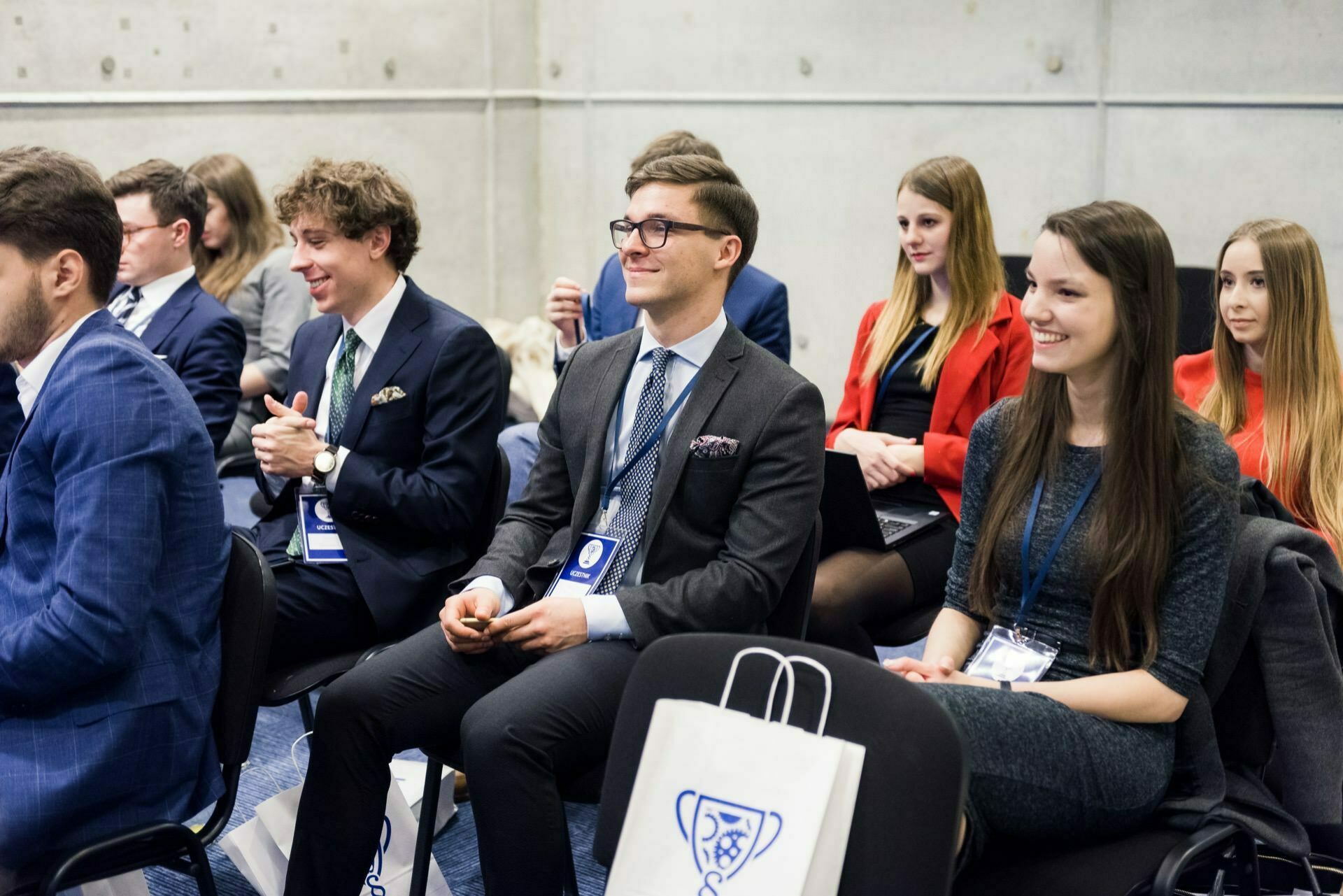 A group of people in business attire sit in rows of chairs in a conference room. Some are smiling and clapping. They have conference badges and bags with logos, perfectly capturing the essence of a professional event. This excellent photo report of the event shows the positive atmosphere against the concrete wall.   