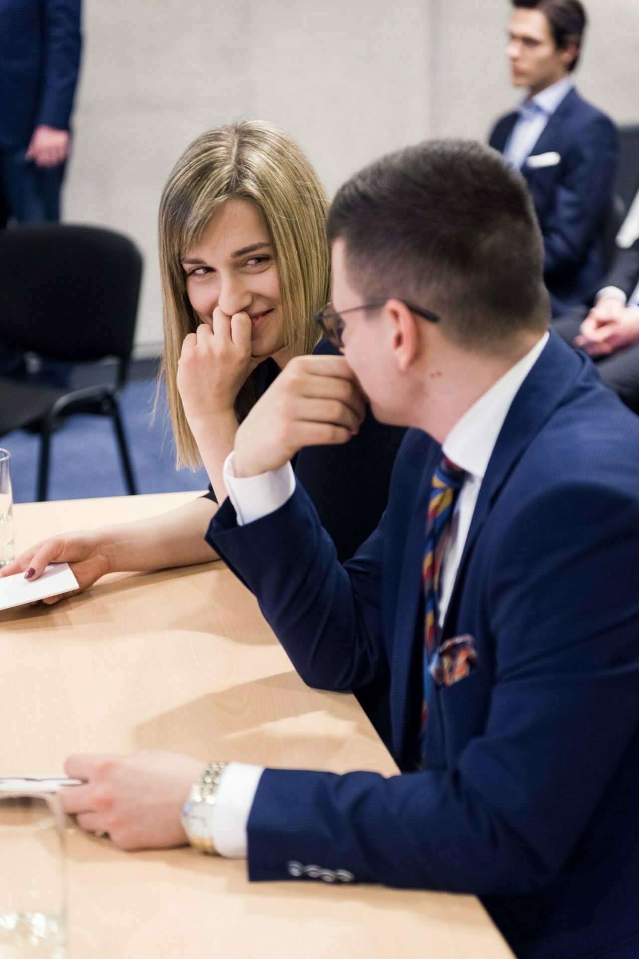 The woman looks at the man sitting next to her at the table with a playful smile. Both are dressed in formal attire and appear to be engrossed in a private conversation, which this event photo captures perfectly. Other seated people are a bit out of focus in the background.  