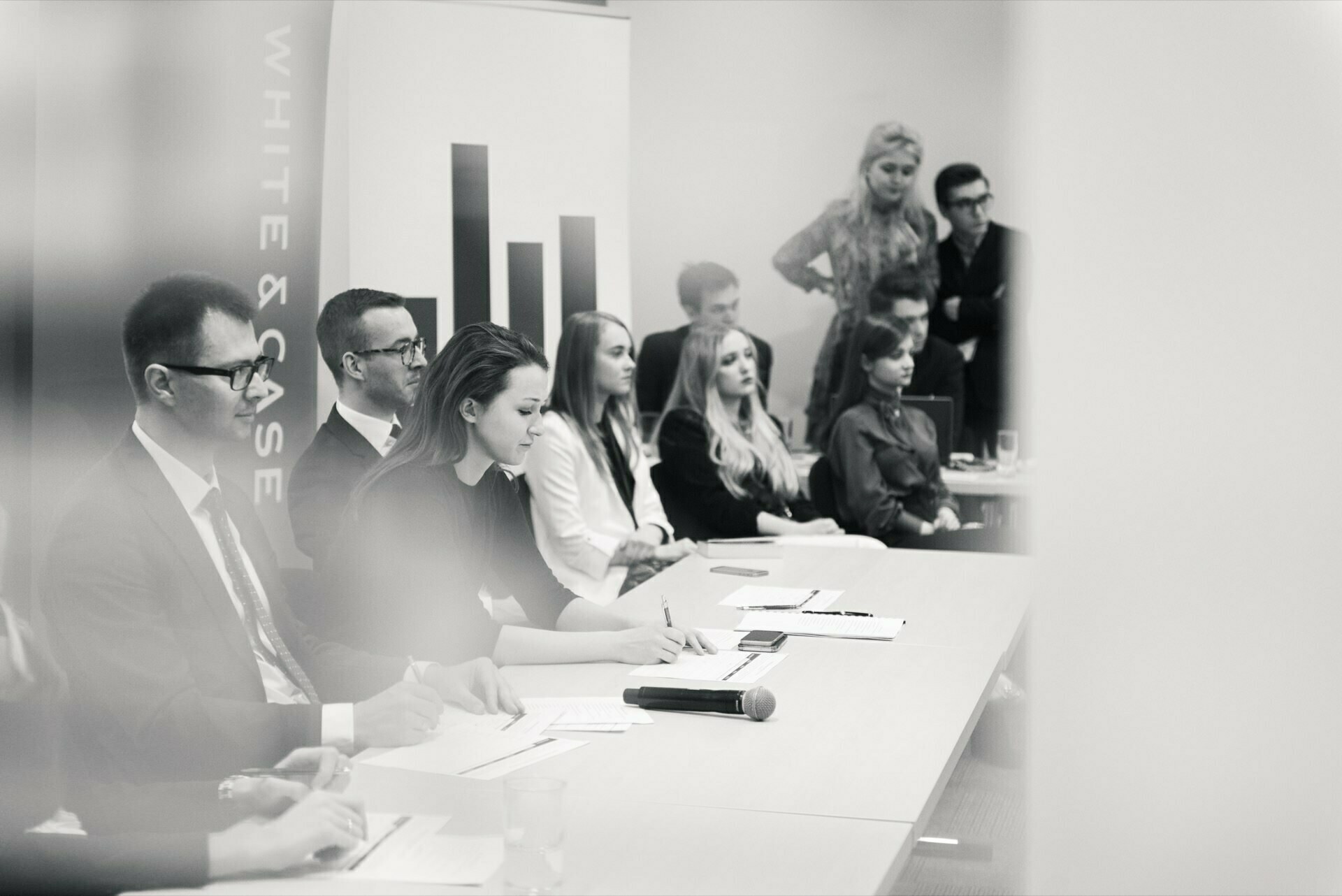 A group of people dressed in business attire sitting at a conference table with notepads and papers. A microphone lies on the table. Some people stand and watch in the background. A vertical banner in the background reads "WHITE AND CASE." This perfectly captures the event photo coverage by a talented event photographer in Warsaw.    