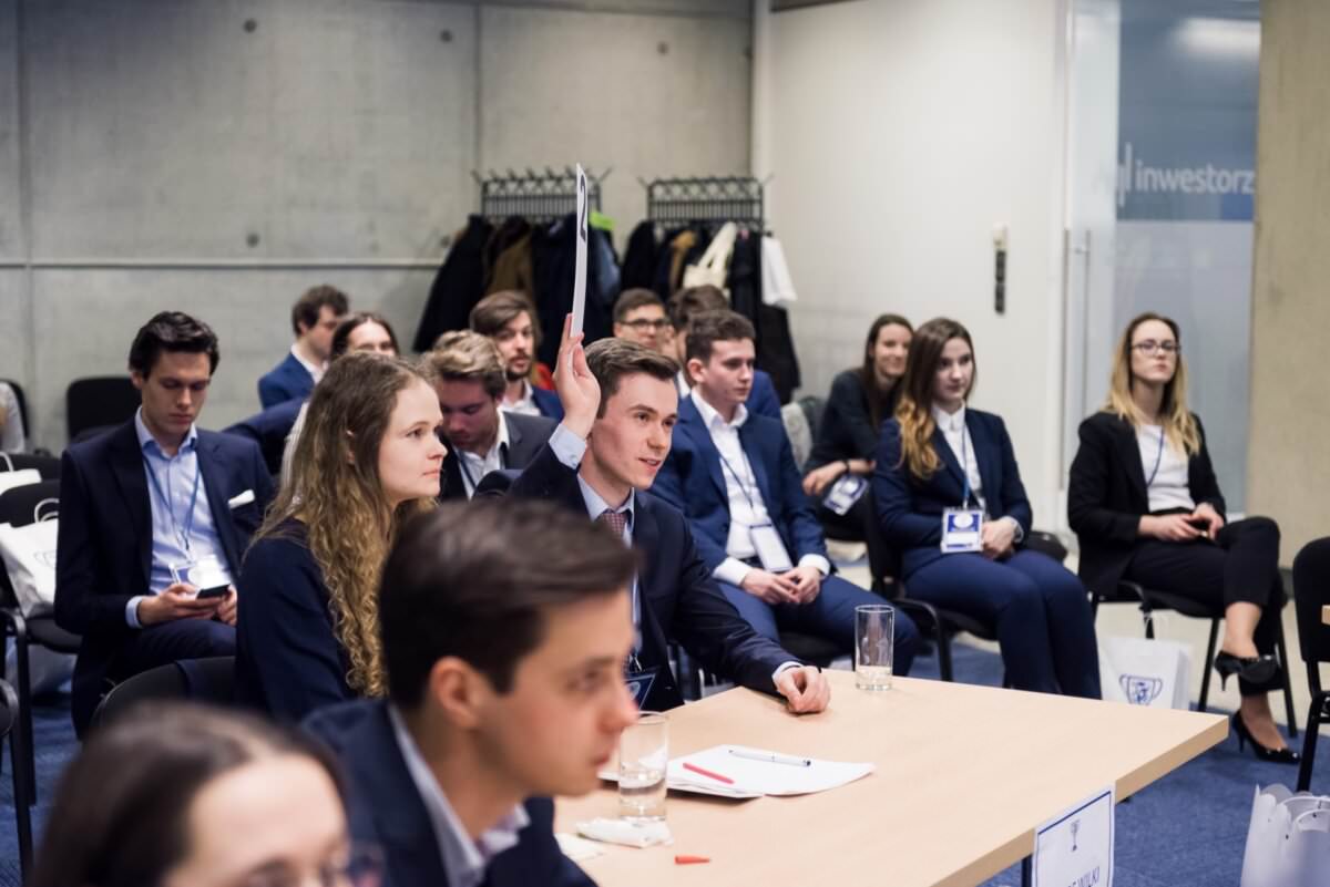 Eine Gruppe von professionell gekleideten Menschen sitzt aufmerksam in einem Konferenzraum. Einige machen sich Notizen, andere hören aufmerksam zu. Im Vordergrund spricht ein Mann. Die Mäntel und Taschen, die auf einem Kleiderbügel im Hintergrund zu sehen sind, fangen die Essenz des professionellen Event-Fotojournalismus in einer geschäftlichen Atmosphäre ein.