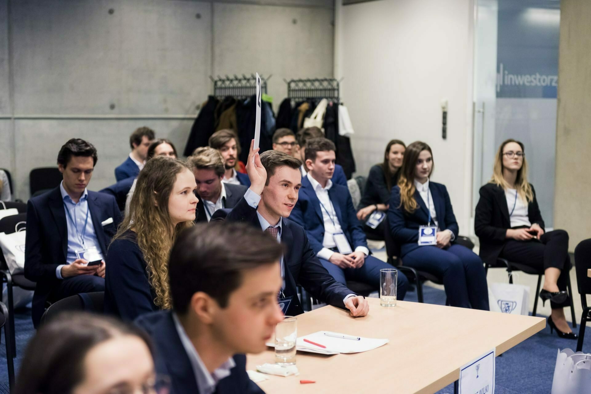 A group of professionally dressed people sit attentively in a conference room. Some are taking notes, others are listening intently. A man is speaking in the foreground. Coats and bags visible on a hanger in the background capture the essence of professional photo coverage of events in a business atmosphere.   