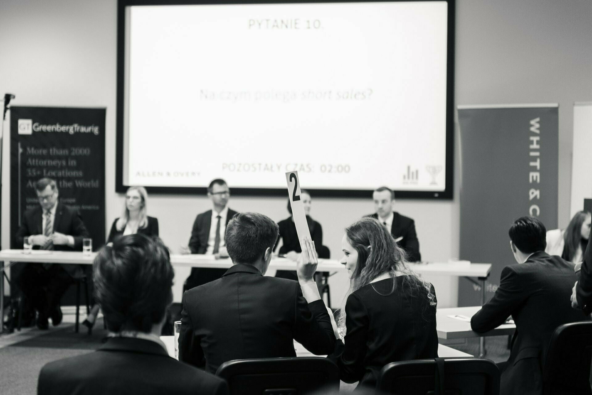 The black and white image shows a formal conference or quiz. Several people are seated at a table on the stage, facing the audience. One person holds a tablet with the number "2" on it. A screen in the background displays a question in Polish, epitomizing classic event photography.   