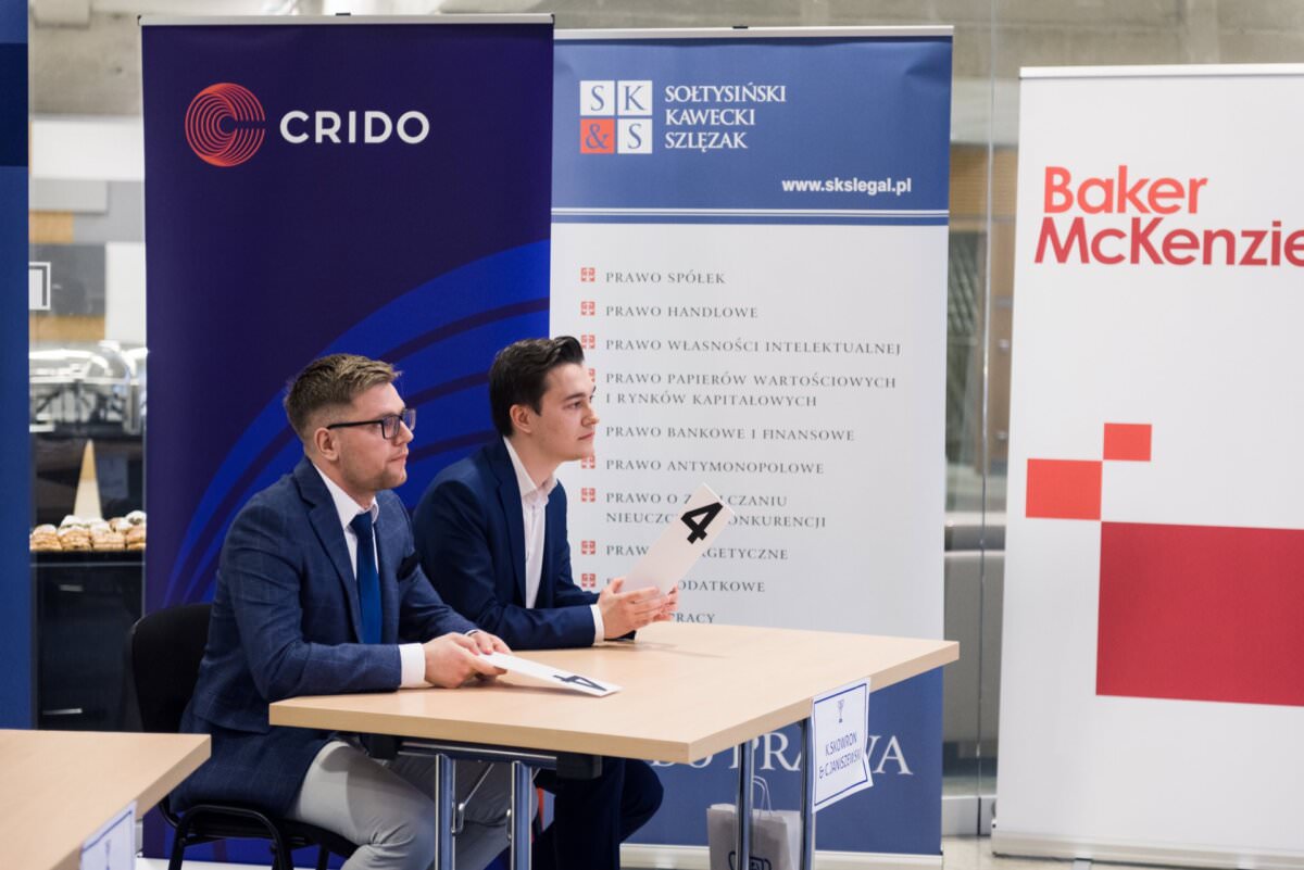 Zwei Männer sitzen an einem Tisch vor Bannern mit Logos und Firmennamen: CRIDO, Soltysinski Kawecki & Szlęzak und Baker McKenzie. Sie sehen aus, als wären sie auf einer professionellen Veranstaltung oder einem Meeting, und einer von ihnen hält ein Mikrofon in der Hand - der perfekte Ort für Eventfotografie.