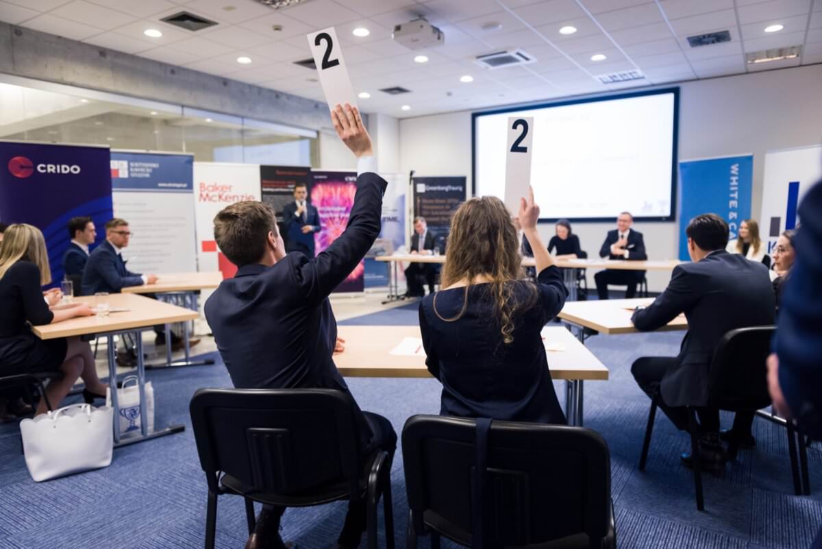 In einem modernen Konferenzraum mit mehreren Tischen und Präsentationsbildschirmen nehmen die Teilnehmer in Geschäftskleidung ihre Nummernkarten in die Hand, während sie diskutieren. Der Raum ist gut beleuchtet und im Hintergrund sind prominente Firmenbanner zu sehen, die die Essenz der Eventfotografie durch einen talentierten Eventfotografen Warschau einfangen.