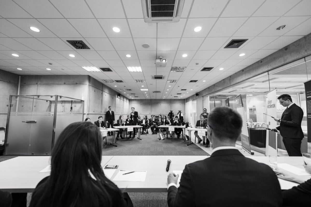 Schwarz-Weiß-Foto, das eine formelle Sitzung in einem Konferenzraum zeigt. Ein Redner steht auf der rechten Seite und wendet sich an das Publikum, während zwei Personen mit dem Rücken zur Kamera an einem Tisch sitzen und auf die in Reihen aufgereihte Gruppe blicken. Diese faszinierende Eventfotografie fängt die Essenz der Veranstaltung perfekt ein.