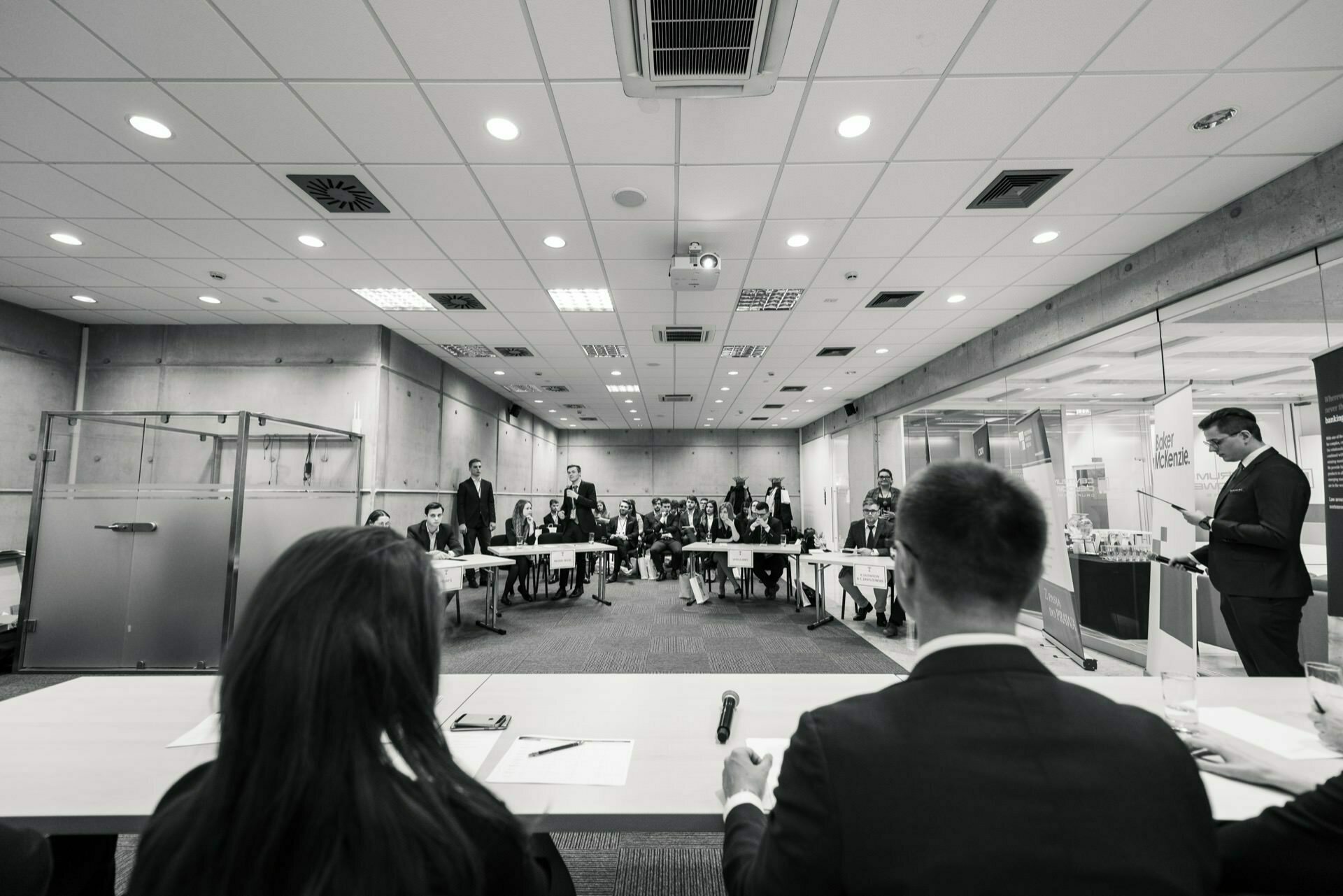 Black and white photo showing a formal meeting in a conference room. A speaker stands on the right, addressing the audience, while two people with their backs turned to the camera sit at a table, facing the group lined up in rows. This fascinating event photography perfectly captures the essence of the event.  