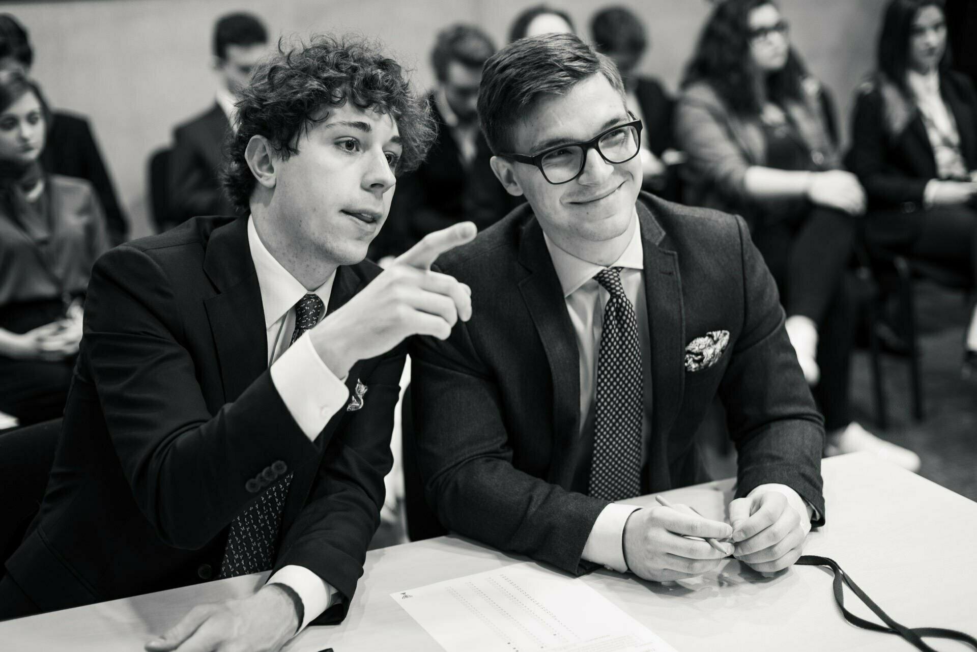 Black and white image showing two young men in suits sitting at a table. One man with curly hair points forward, while the other, wearing glasses and smiling, holds a pen. Several people in business attire are sitting in the background, which is perfectly captured in an event photo by event photographer Warsaw.  