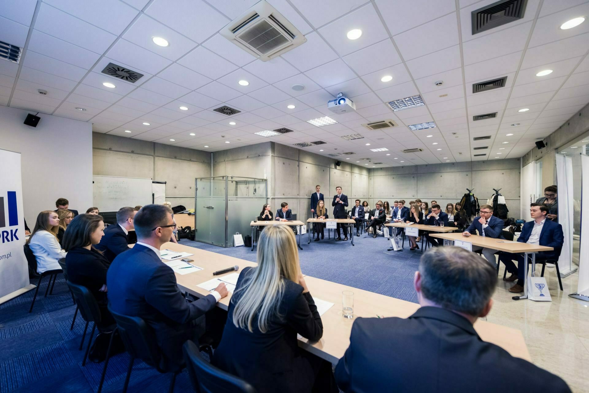 A group of people in business attire sit around a large U-shaped table in a modern conference room. Some are listening intently, others are taking notes. The room, which resembles a typical setting from "event photography," features a whiteboard, glass partitions and a projector screen. A banner with the word "WORK" is partially visible on the left.   