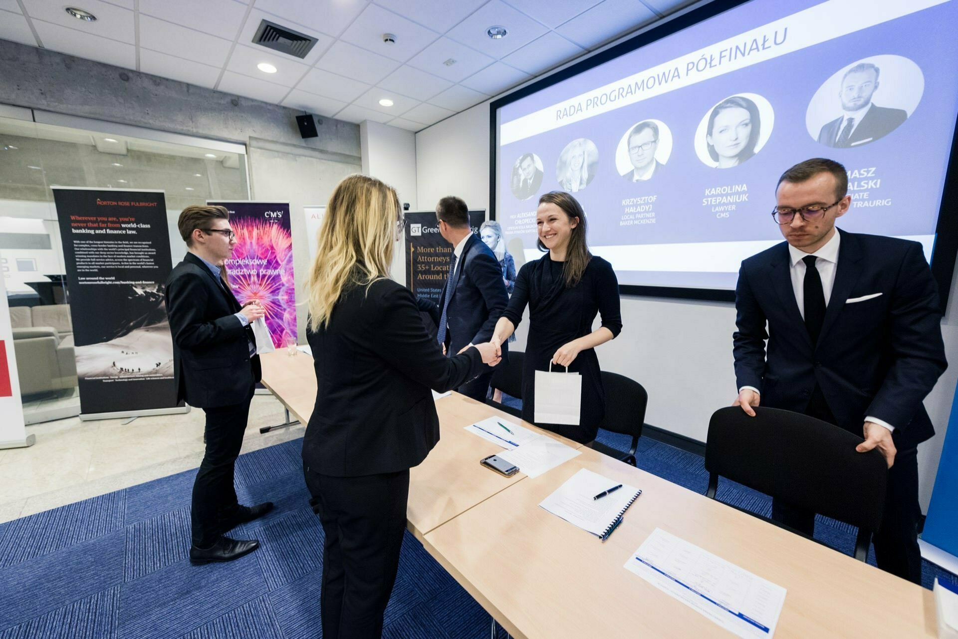 A group of professionals is in a conference room. Two people are shaking hands at a table while others stand and talk. Behind them, a large screen displays profiles of people with text in another language. The setting takes on a formal character, capturing the essence of event photography.   