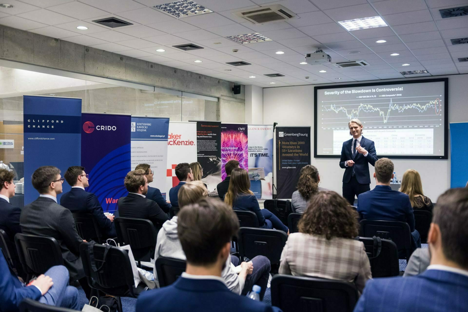 A speaker in a suit addresses an audience in a conference room. Attendees sit facing forward and look at a projector screen displaying charts and graphs. At the front of the room, next to the speaker, numerous banners of various sponsors are placed, capturing the essence of event photography.  