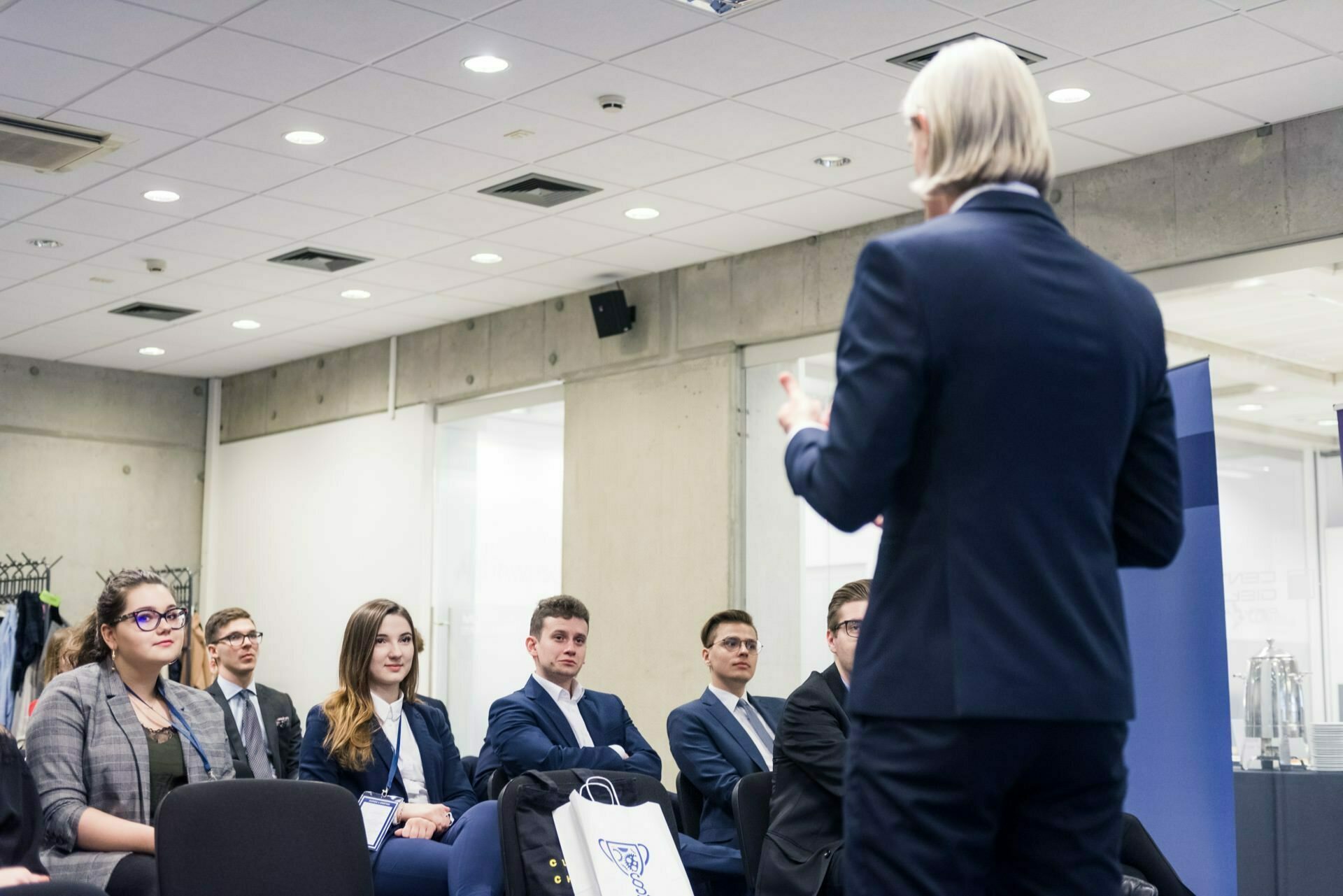 A person with long, light hair and wearing a suit is giving a presentation in an office. In front of the speaker sit several attendees, both men and women, dressed in business attire, listening intently. This moment captures the essence of event photography at its best.  