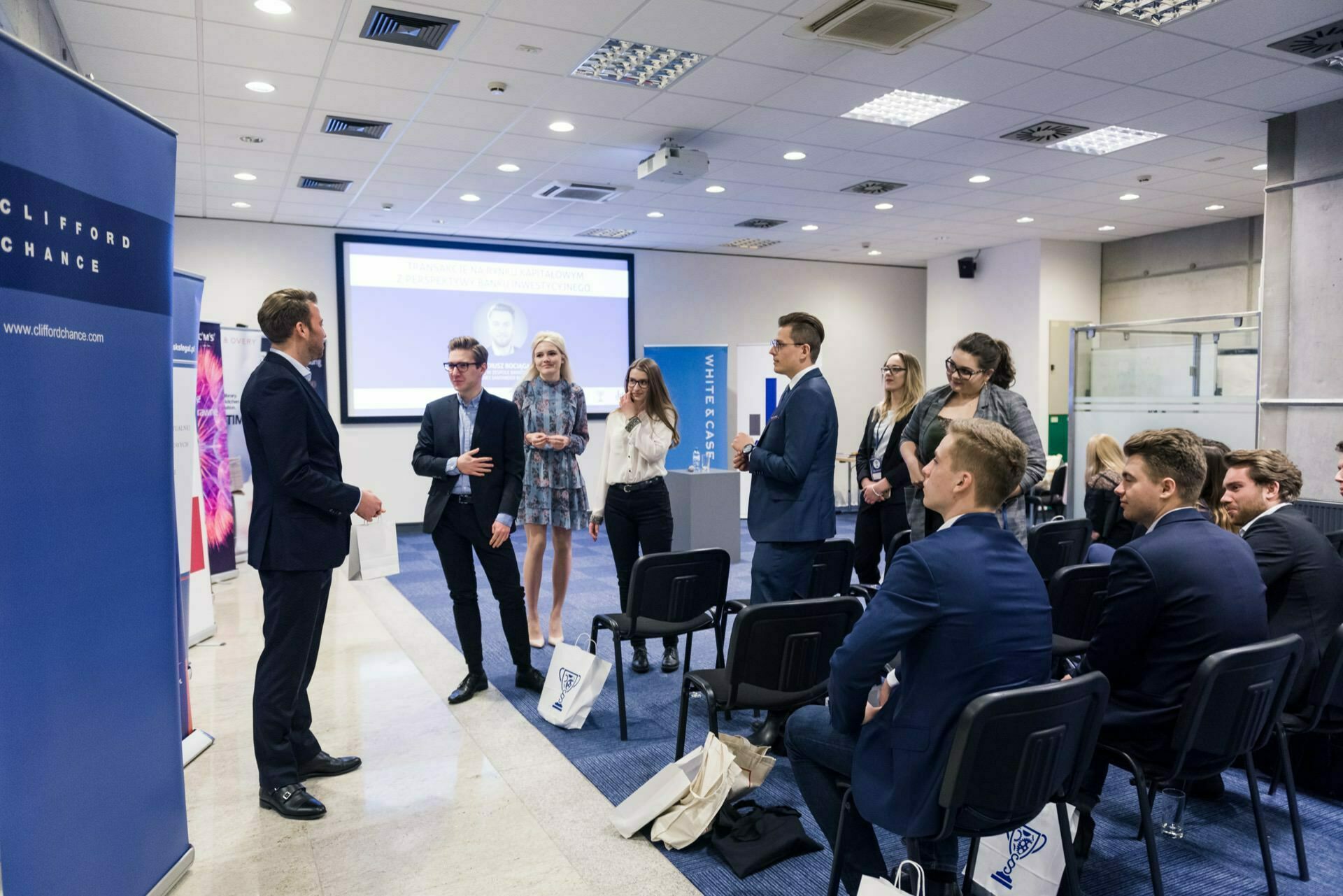 A man in a suit stands and speaks to a group of young professionals in a conference room. The group, dressed in business attire, listens attentively while standing next to rows of chairs. In the background, screens and banners are visible, capturing the essence of the photo event.  