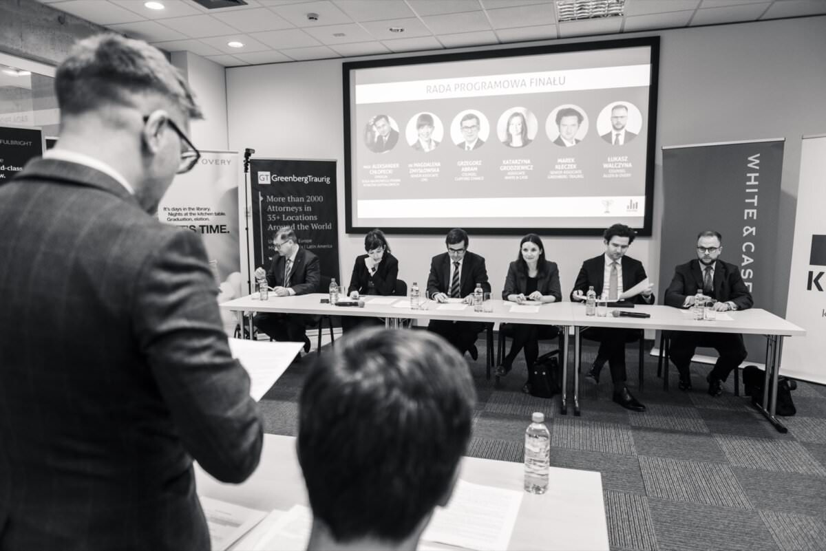 Schwarz-Weiß-Bild eines Geschäftstreffens in einem Konferenzraum. Eine Person, die von hinten gesehen wird, steht und liest ein Dokument im Vordergrund. Sechs Personen sitzen an einem langen Tisch und hinter ihnen befindet sich eine Projektionsfläche, auf der Namen und Fotos zu sehen sind - eine ideale Fotoreportage von Ereignissen, die berufliche Interaktionen zeigen.