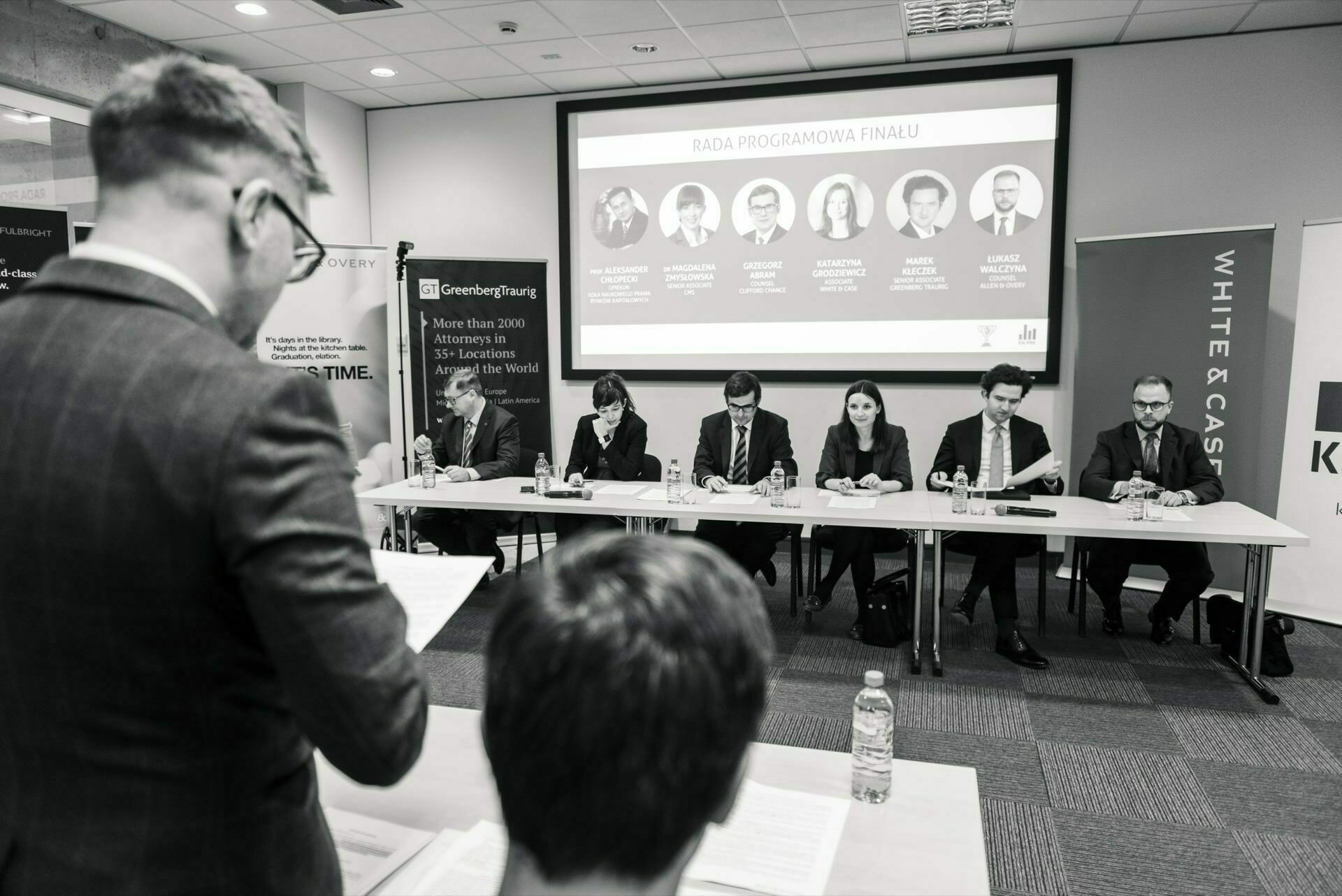 Black and white image of a business meeting in a conference room. A person seen from behind stands and reads a document in the foreground. Six people are seated at a long table, and behind them is a projection screen displaying names and photos - an ideal photo-report of events showing professional interactions.  