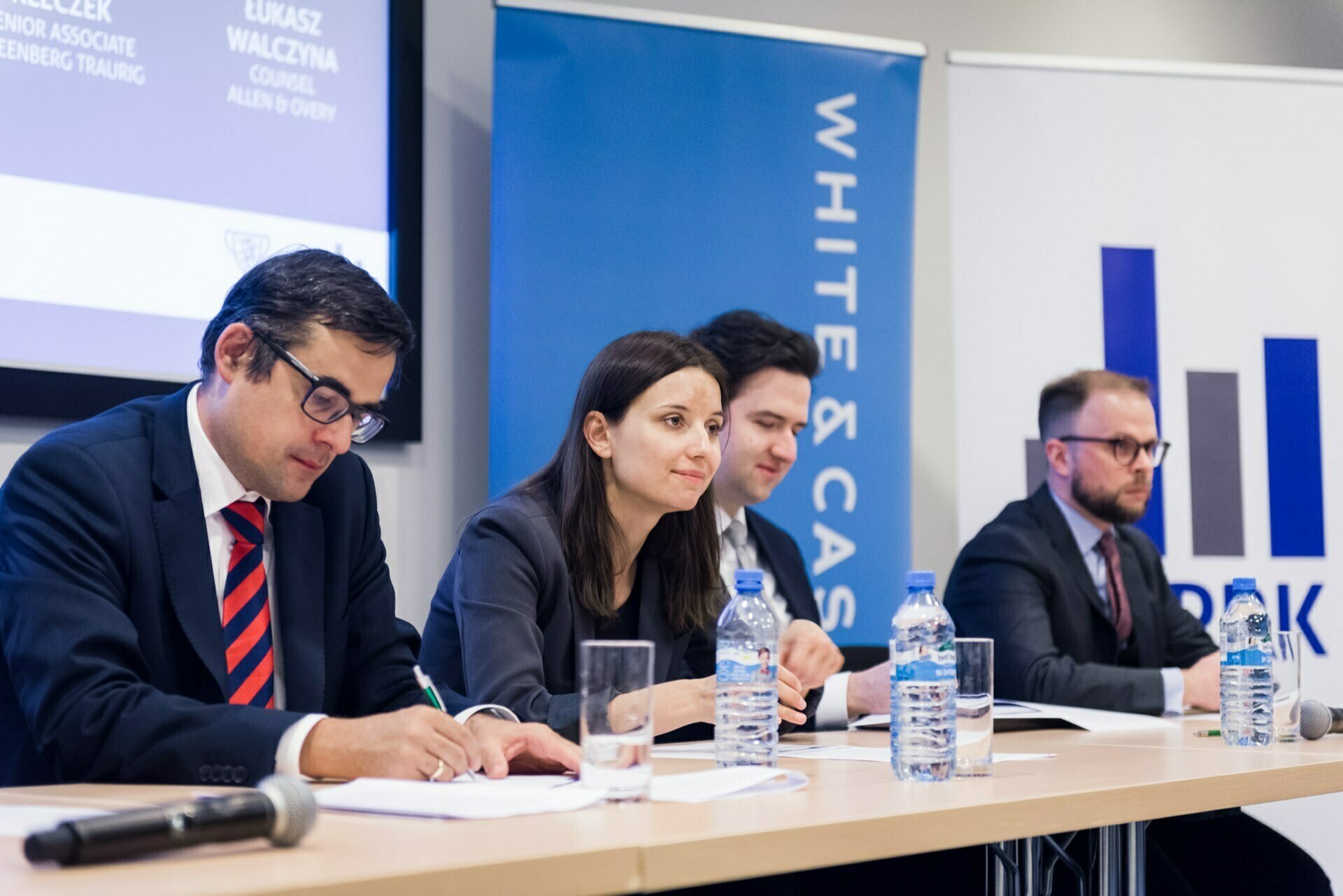 Four people sit at a conference table, each equipped with a microphone and water bottles, and participate in a panel discussion. Behind them are two banners with the words "White &amp; Case" and "RAK". Event photographer warsaw expertly captured this event photo essay.  