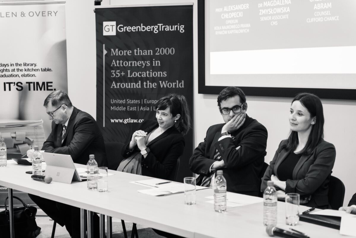 Das Schwarz-Weiß-Foto zeigt vier Fachleute, die während eines Fototermins der Veranstaltungen an einem Diskussionstisch sitzen. Hinter ihnen ist ein Banner mit der Aufschrift 'Greenberg Traurig' und Angaben zu ihrer weltweiten Präsenz zu sehen. Auf dem Tisch stehen Wasserflaschen und Mikrofone, und einer der Diskussionsteilnehmer arbeitet mit einem Laptop.