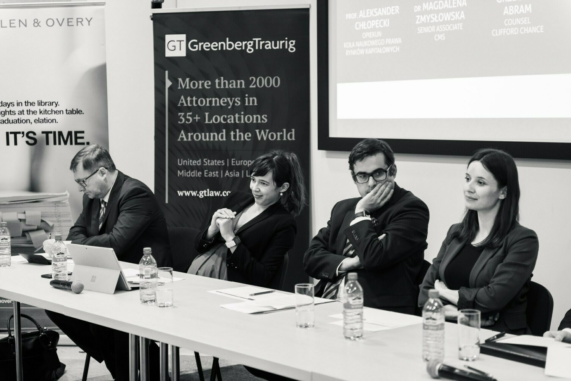 The black-and-white photo shows four professionals sitting at a discussion table during a photo-op of the events. Behind them is a banner with the words "Greenberg Traurig" and details of their global presence. Water bottles and microphones stand on the table, and one of the panelists is using a laptop.  