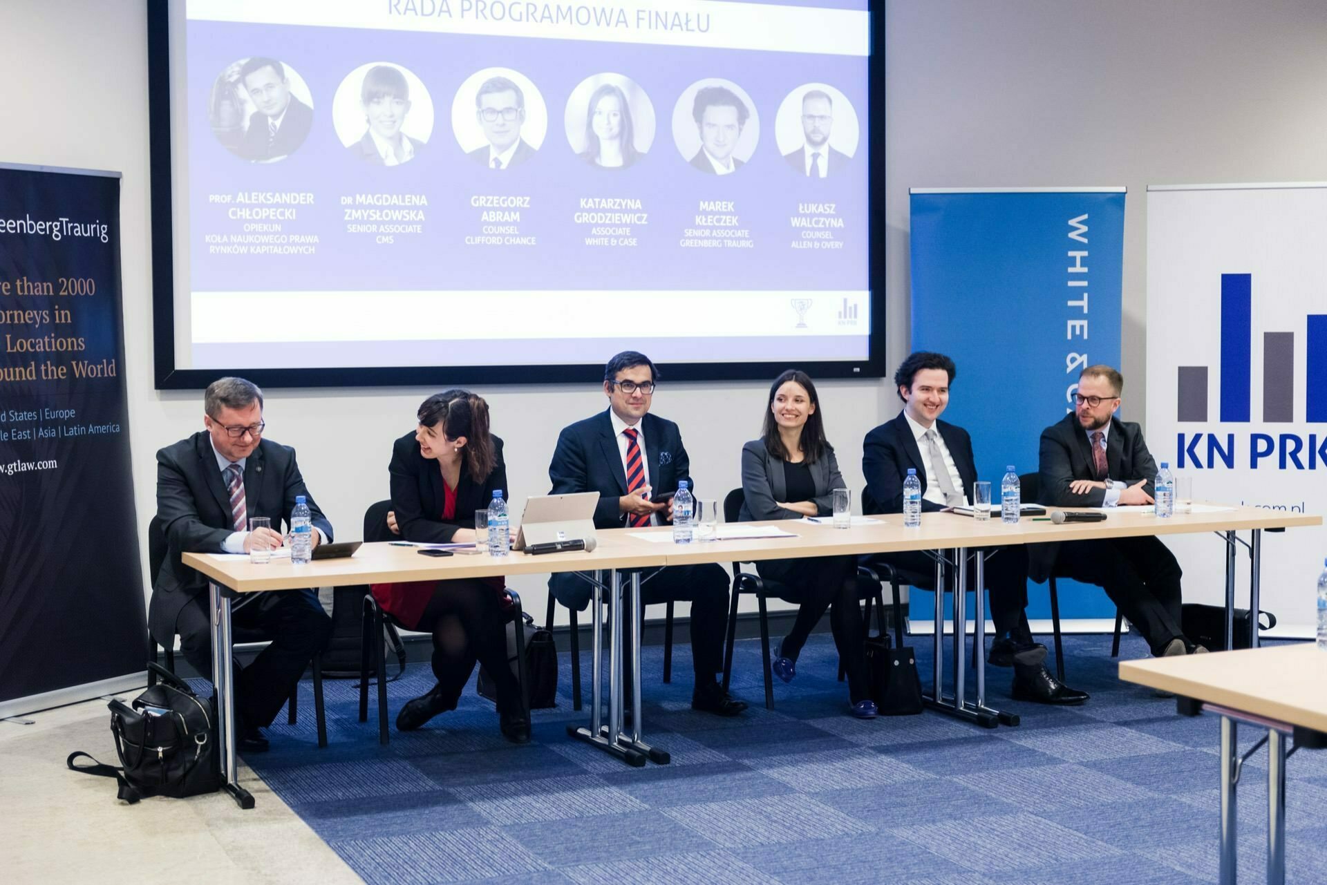 A group of six people sit at a long table in a conference room. Behind them, a screen displays information about the event and their names. Greenberg Traurig and KN Przy banners are visible on both sides of the room. The atmosphere is professional and formal, perfect for event photography by event photographer Warsaw.   