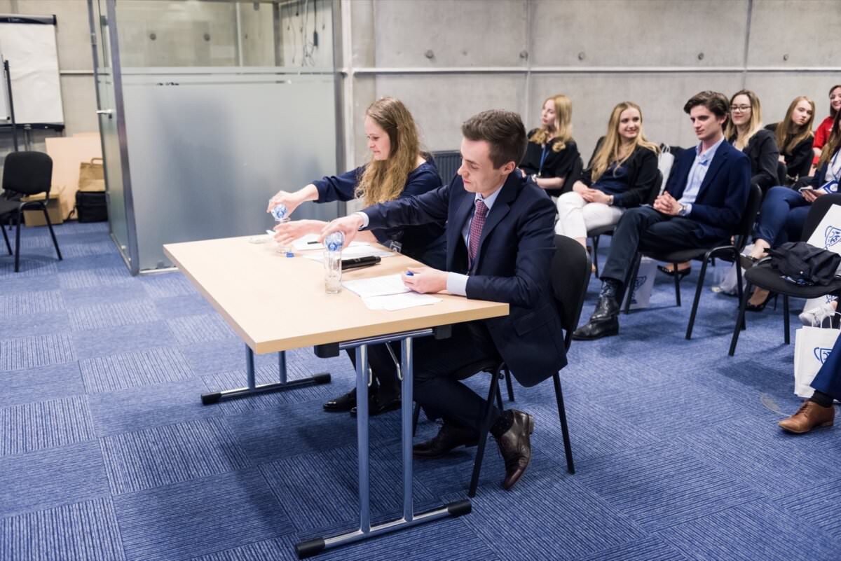 In dem Raum sitzt eine Gruppe junger Erwachsener in Business-Kleidung. Zwei Personen am vorderen Tisch schütteln Hände über Dokumente und lächeln. Andere im Hintergrund scheinen beschäftigt zu sein, einige sitzen, andere stehen. Die Umgebung lässt auf ein formelles Treffen oder eine Veranstaltung schließen, ideal für Eventfotografie.