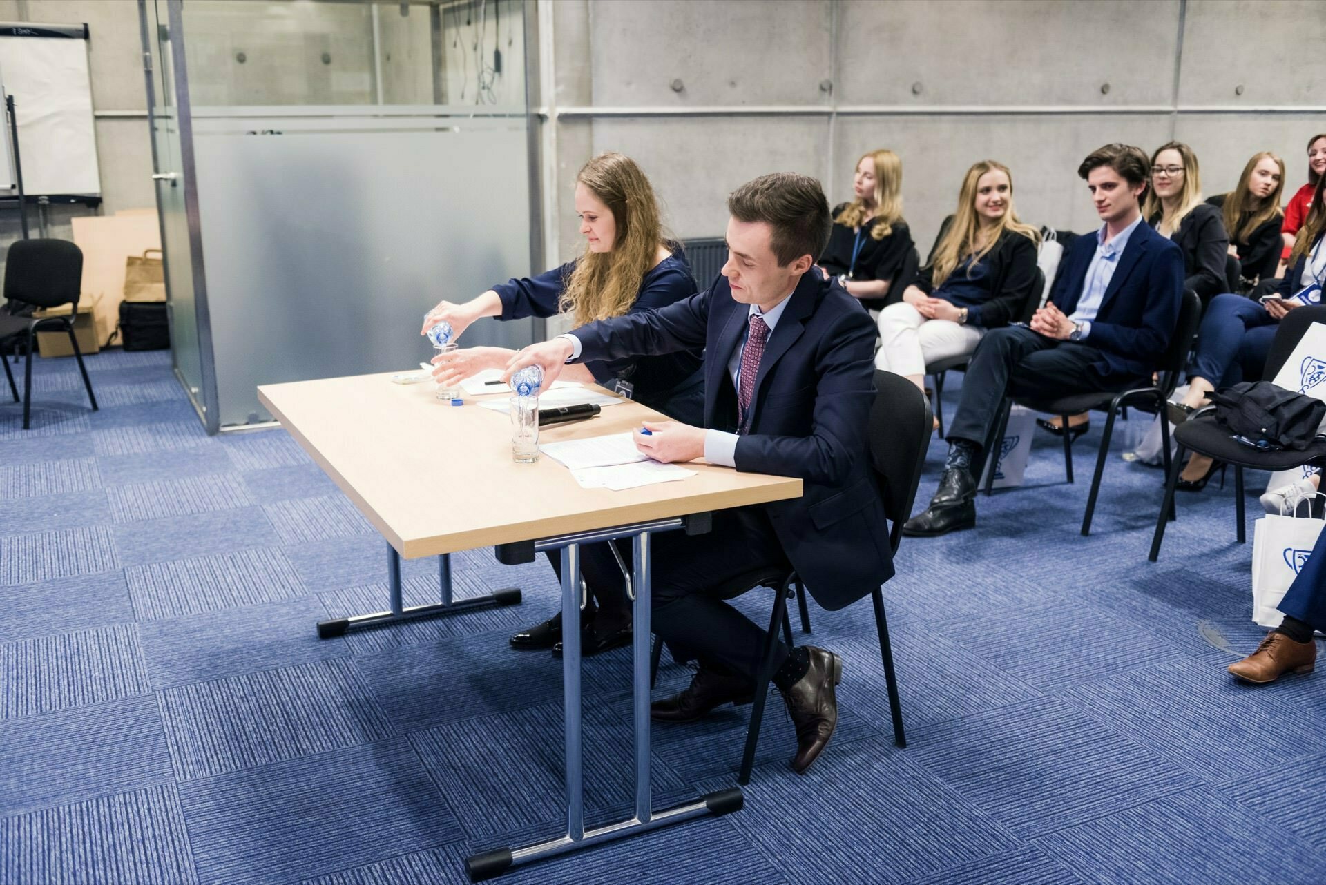 Sitting in the room is a group of young adults dressed in business attire. Two people at the front table are shaking hands over documents and smiling. Others in the background appear engaged, some sitting and others standing. The setting suggests a formal meeting or event, perfect for event photography.   