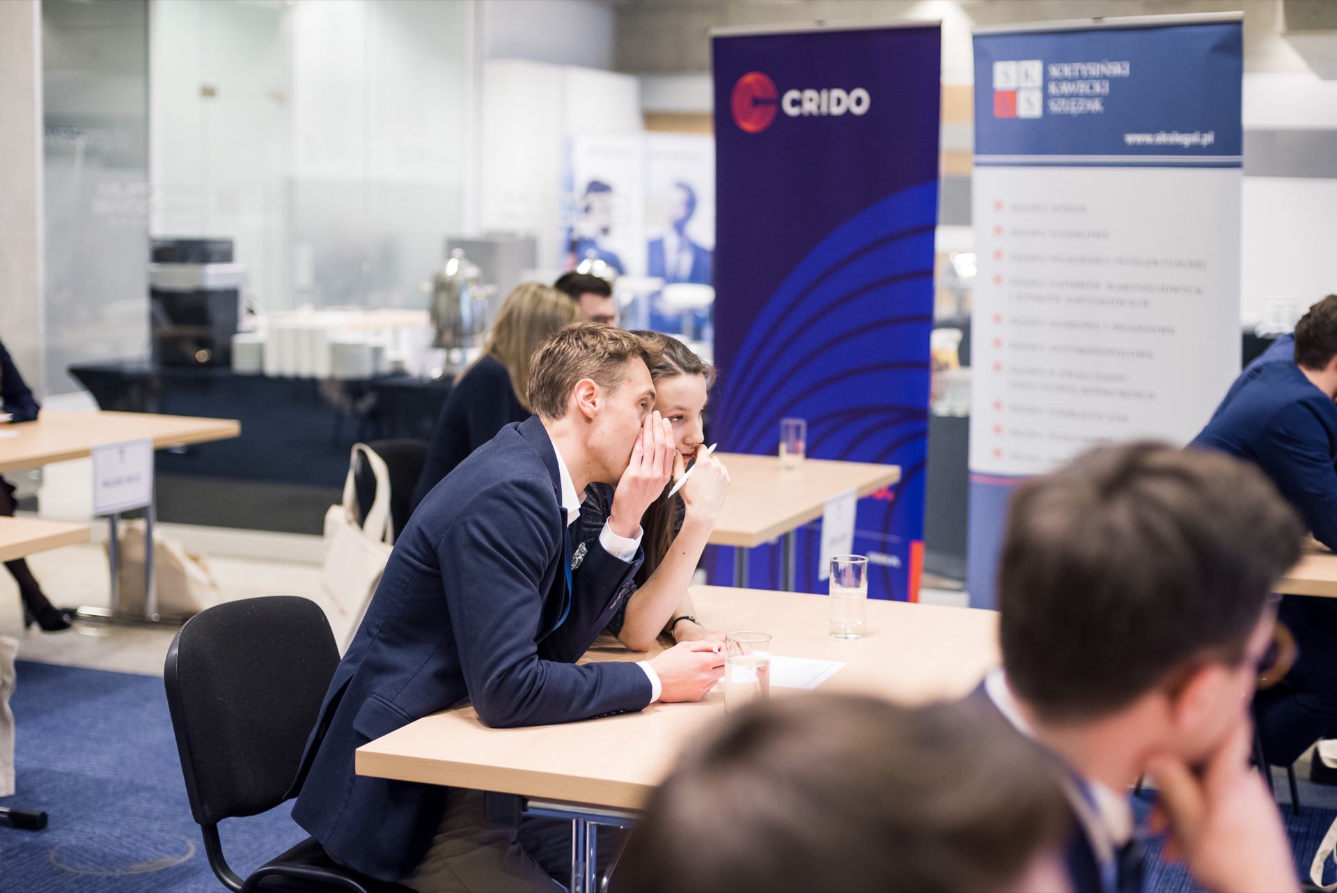 People sitting at tables in a conference setting. The man and woman in the center are talking to each other, while other participants are scattered around the room. In the background are two large branded banners with the words "CRIDO" and "SIX". This could be an ideal scene for event photography or photojournalism.   