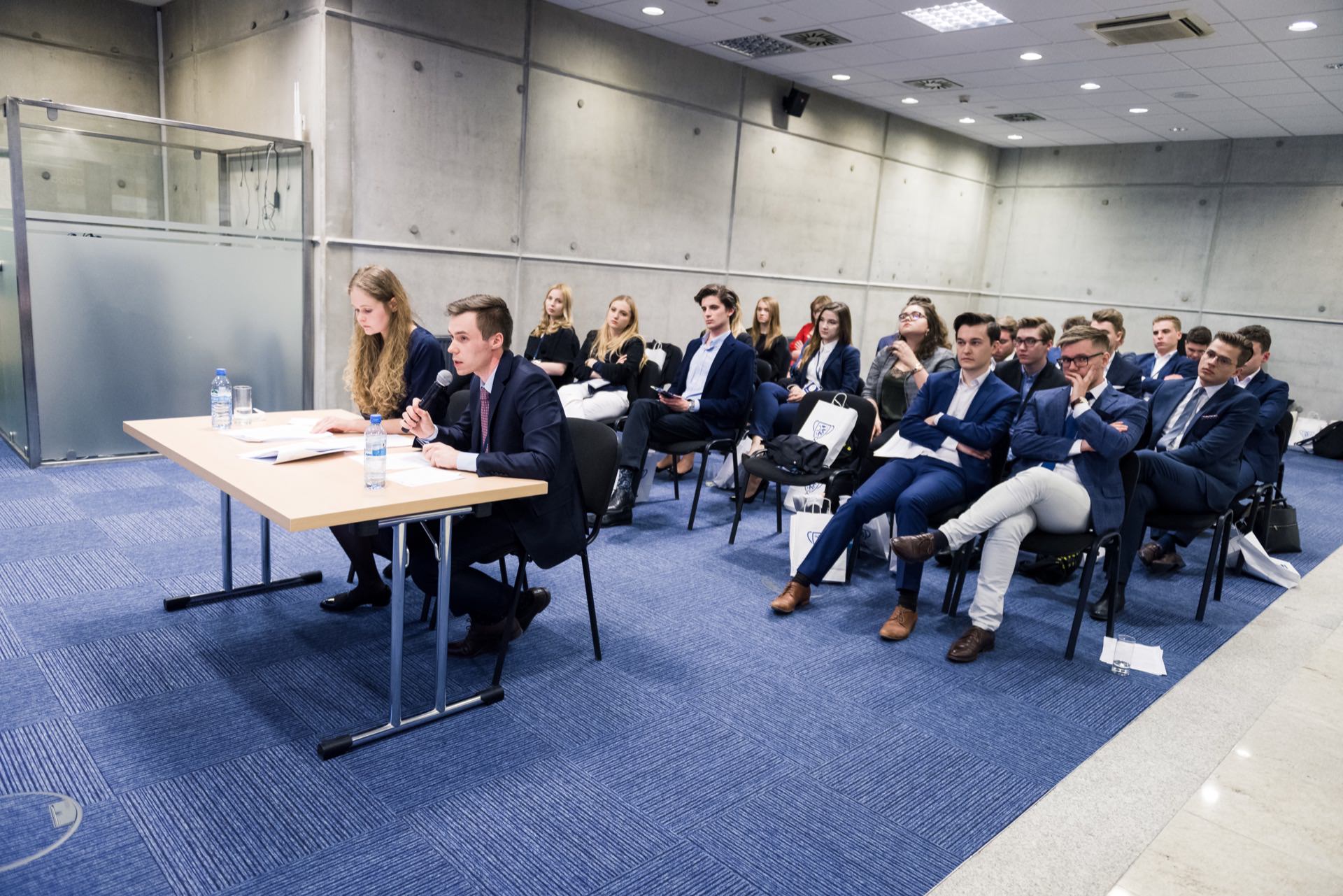 A group of people in business attire attend a presentation in a conference room. Two people in the front speak to the audience while the participants listen attentively. The room has gray walls and a blue carpet, perfect for an event photography session from event photographer Warsaw.  