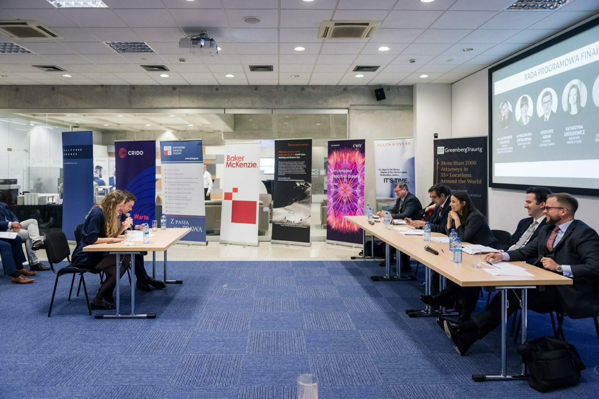 A group of people in professional attire sit at tables in a modern conference room. In the background hang banners of various organizations and a large screen displaying information. In the foreground, two people are sitting at a table, facing each other, which is perfectly captured by the event photographer warsaw.  