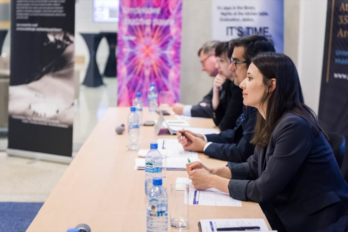 Eine Gruppe von Personen in Geschäftskleidung sitzt an einem langen Tisch, hört aufmerksam zu und macht sich während einer Konferenz Notizen. Wasserflaschen, Notizbücher und Stifte liegen auf dem Tisch. Auf Bannern im Hintergrund sind verschiedene Grafiken und Texte zu sehen, die sich auf die Veranstaltung beziehen. Diese Szene ist ein hervorragendes Beispiel für Eventfotografie in Warschau.