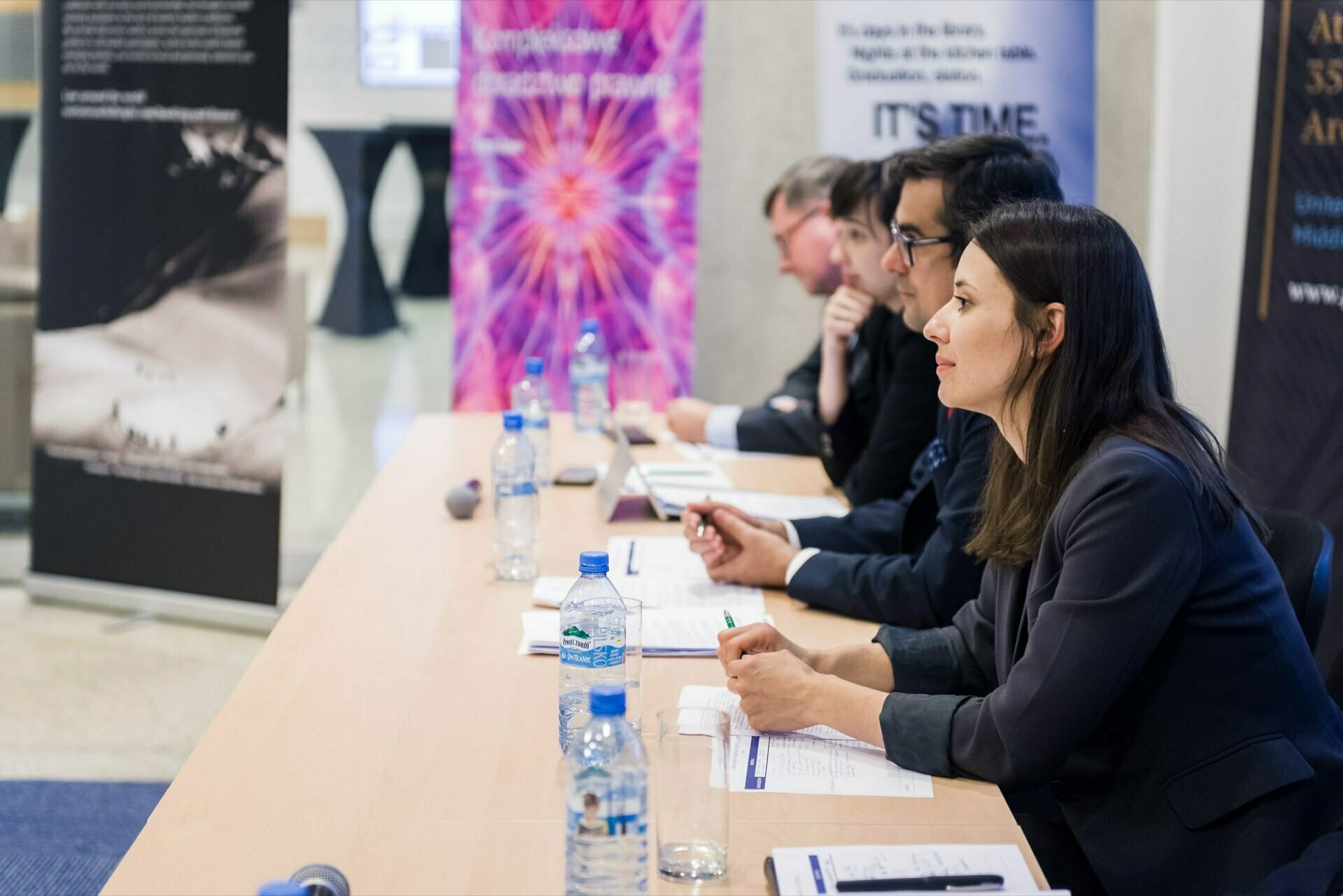 A group of people in business attire sit at a long table, listening intently and taking notes during the conference. Water bottles, notebooks and pens lie on the table. Banners in the background display various graphics and texts related to the event. This scene is an excellent example of event photography in Warsaw.   

