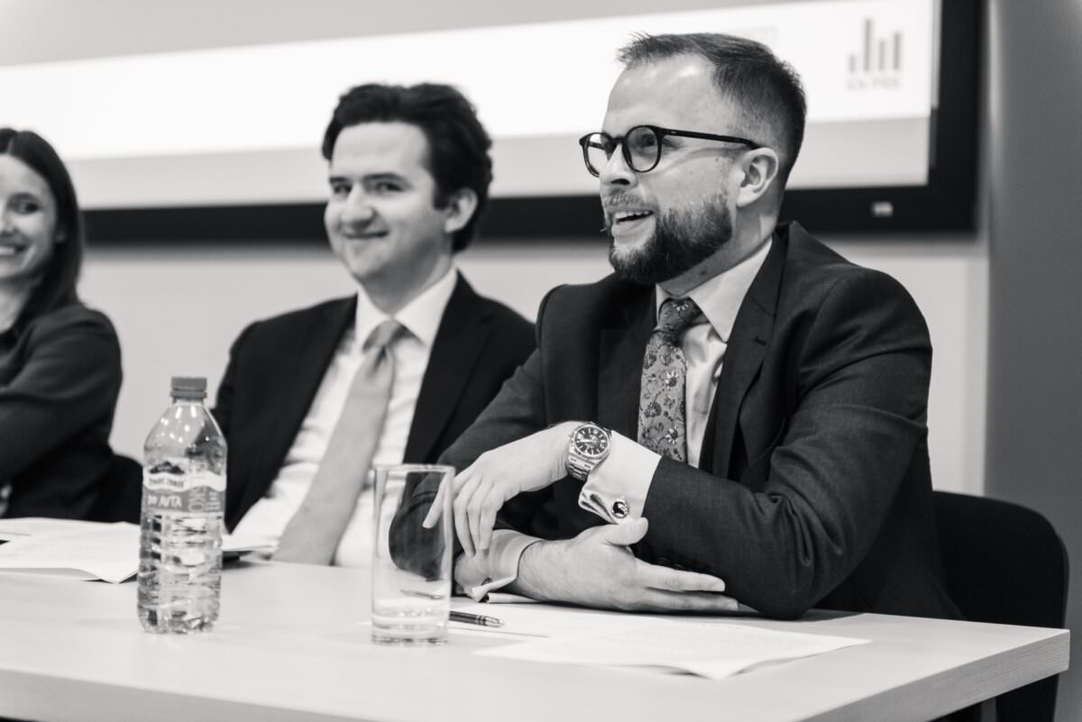 Drei Personen sitzen an einem Konferenztisch, haben Dokumente vor sich liegen und führen eine Diskussion. Der Mann auf der rechten Seite, der eine Brille und einen Anzug trägt, spricht. Eine Frau und ein zweiter Mann, beide ebenfalls im Geschäftsanzug, hören aufmerksam zu. Während dieser Fotosession stehen eine Flasche Wasser und ein Glas auf dem Tisch.