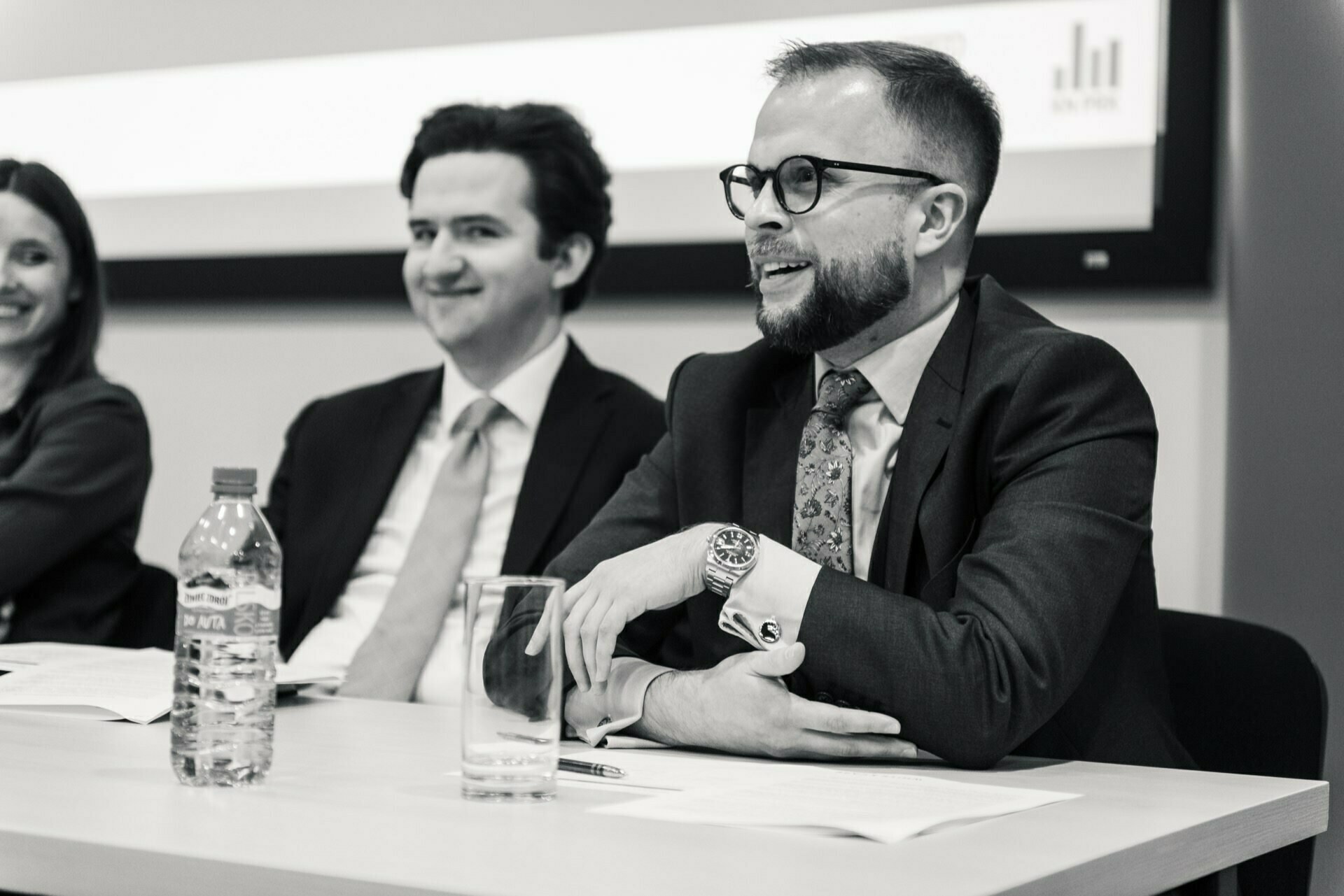 Three people are sitting at a conference table with documents in front of them and having a discussion. The man on the right, wearing glasses and a suit, is speaking. A woman and another man, both also in business attire, are listening attentively. During this photo shoot, a bottle of water and a glass lie on the table.   