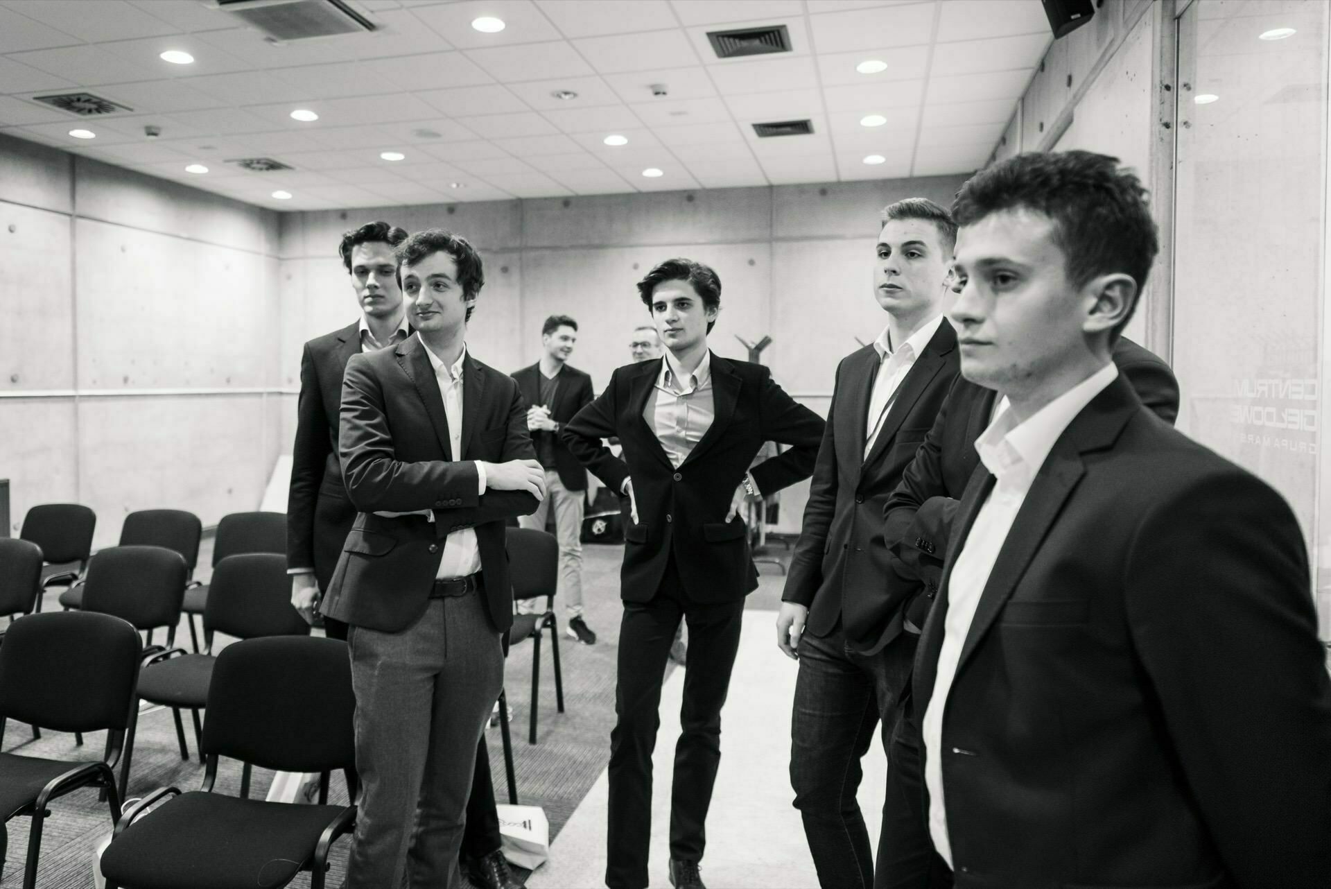 The black-and-white event photograph shows a group of young men in business attire standing in a conference room with rows of empty chairs. Four of the men can be seen in the foreground having a conversation, while the others can be seen in the background, which is an authentic event photo. 