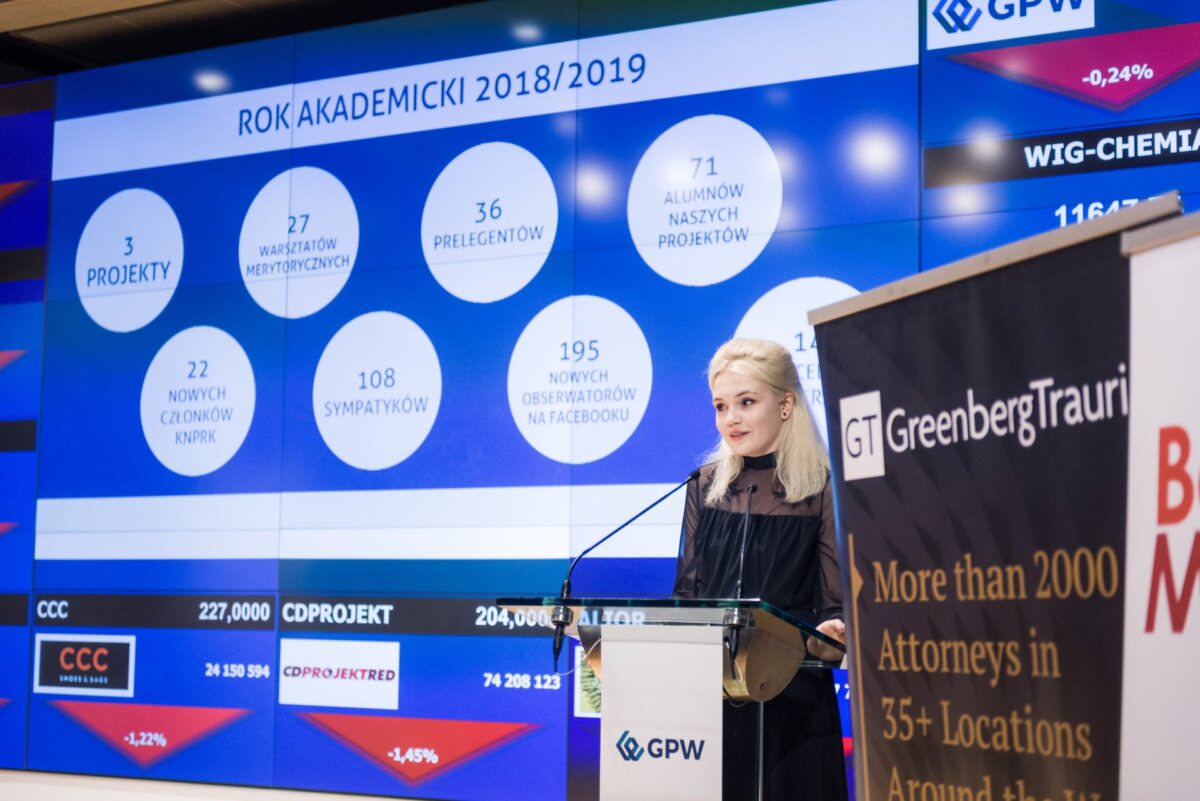 Eine Frau steht an einem Podium und hält eine Präsentation. Hinter ihr befindet sich ein großer Bildschirm, auf dem verschiedene Statistiken auf Polnisch zum akademischen Jahr (2018/2019) angezeigt werden. Neben ihr ist ein Banner mit einem Text über die Anwaltskanzlei Greenberg Traurig zu sehen, perfekt eingefangen in einem Event-Fotoessay des Eventfotografen Warschau.