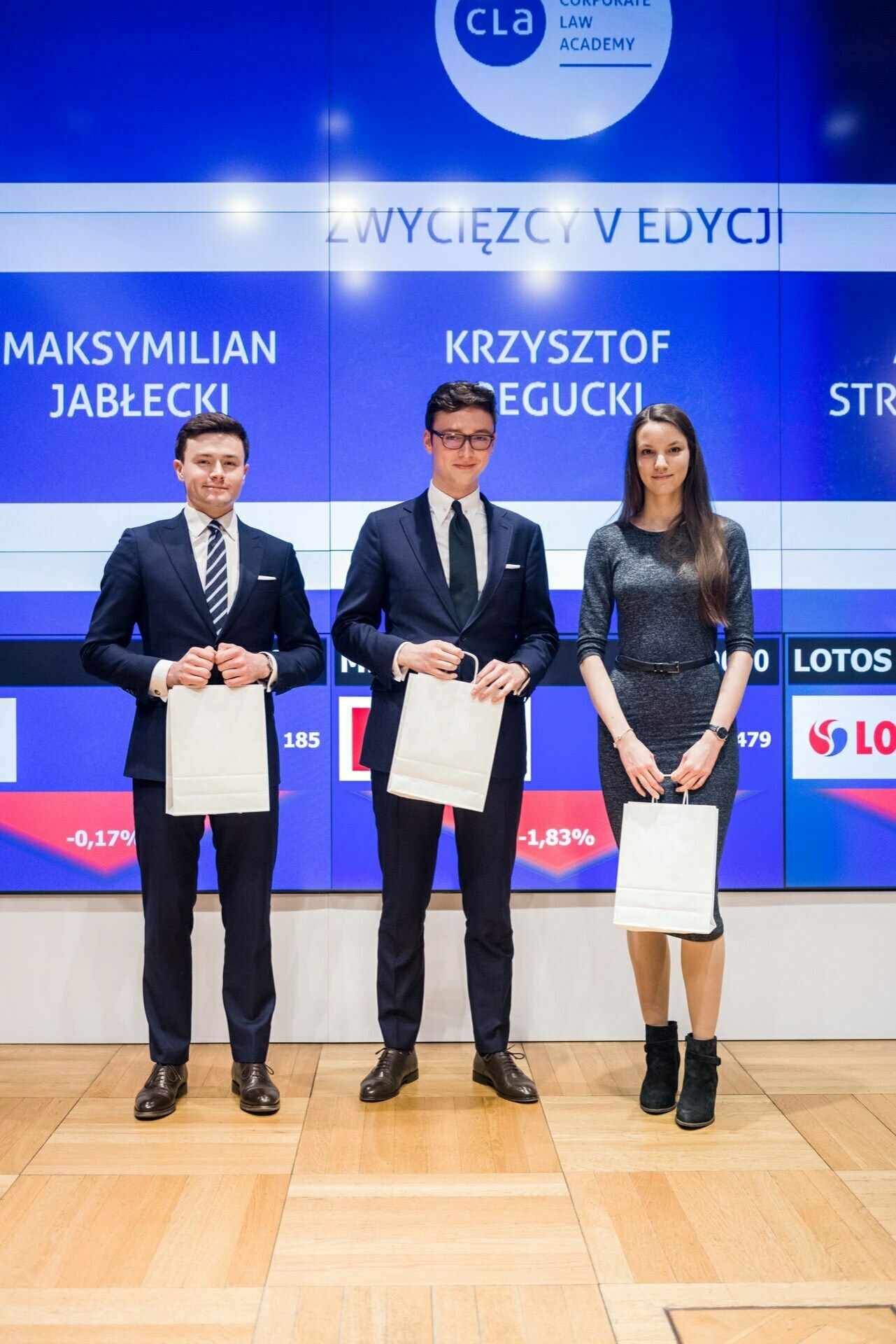 Three people stand in front of a large screen at the event, holding white bags. The two men on the left are wearing suits and ties, while the woman on the right is wearing a matching dress. Their names are projected on the screen behind them - a perfect photo-reportage of the event captured by an event photographer Warsaw.  