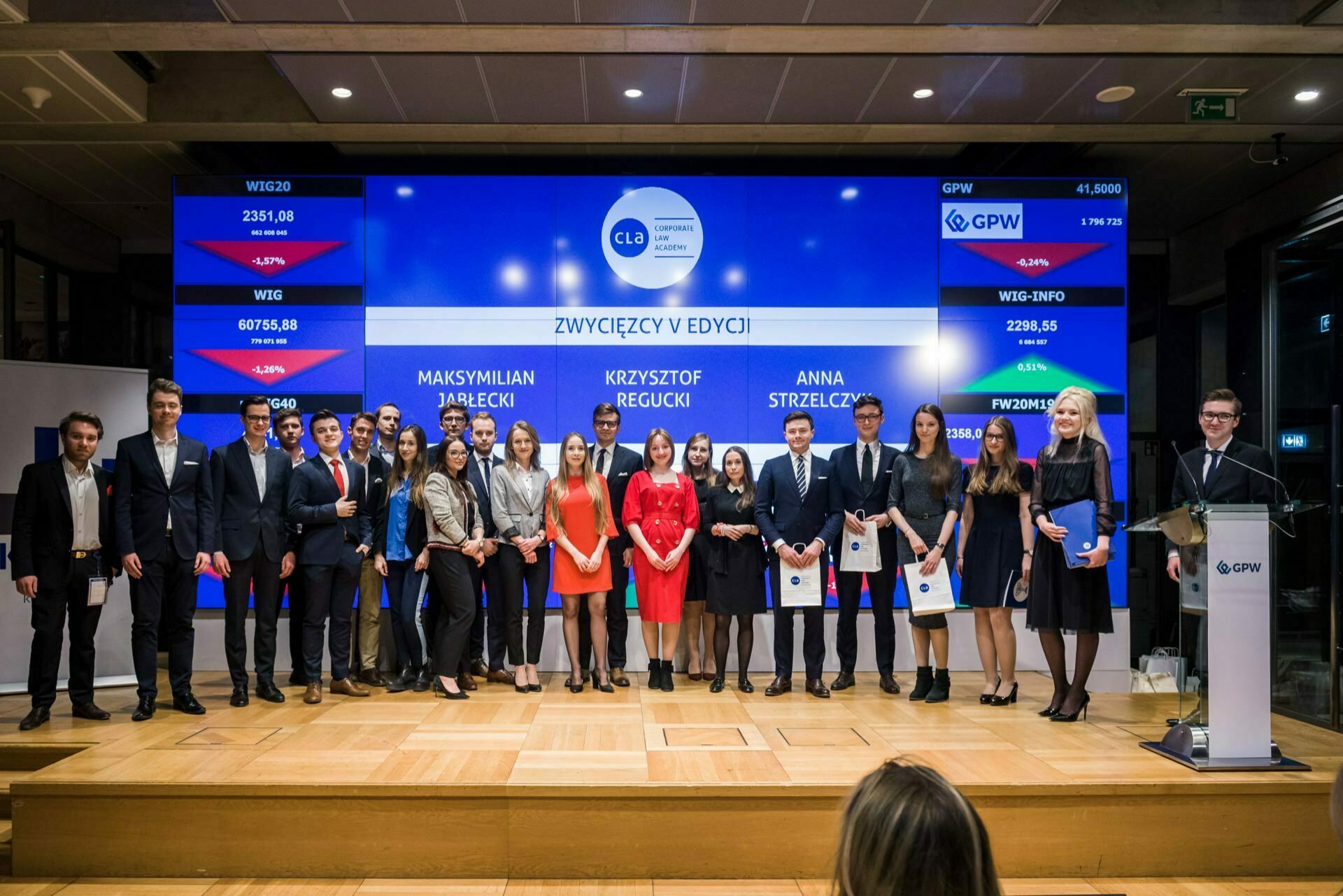 A group of people stand on stage with certificates in front of a large screen displaying names and charts. The screen shows the names of Maksymilian Jarecki, Krzysztof Regucki and Anna Strzelczyk in the background. The formal and ceremonial setting is perfect for event photography.  