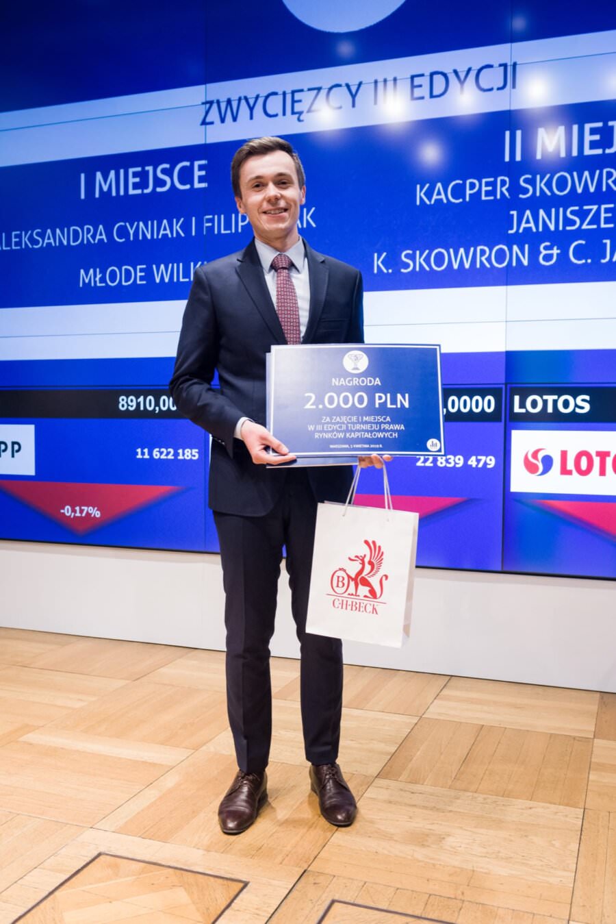 Ein lächelnder Mann im Anzug hält einen großen Scheck über 2.000 £ und eine Geschenktüte vor einem Display, auf dem die Ergebnisse eines Wettbewerbs präsentiert werden. Im Hintergrund sind Firmennamen und Logos in einer professionellen Umgebung zu sehen - ein perfektes Beispiel für Event-Fotojournalismus von einem talentierten Eventfotografen aus Warschau.