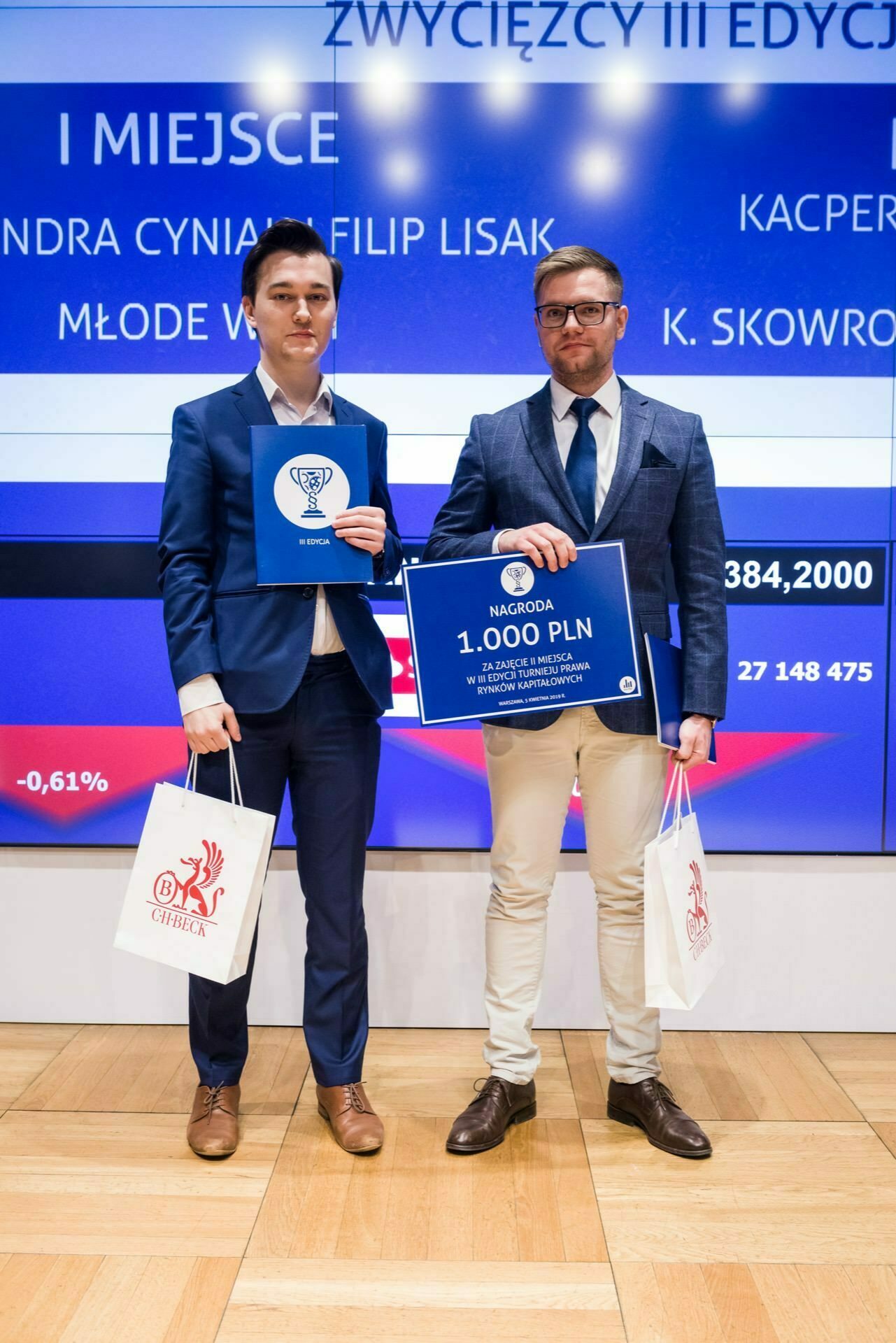 Two men in formal attire stand side by side, holding gift bags - one with a trophy, the other with a large check for $1,000. A digital display in the background indicates that they are at an awards ceremony. This photo is part of an event photo report by event photographer Warsaw.  