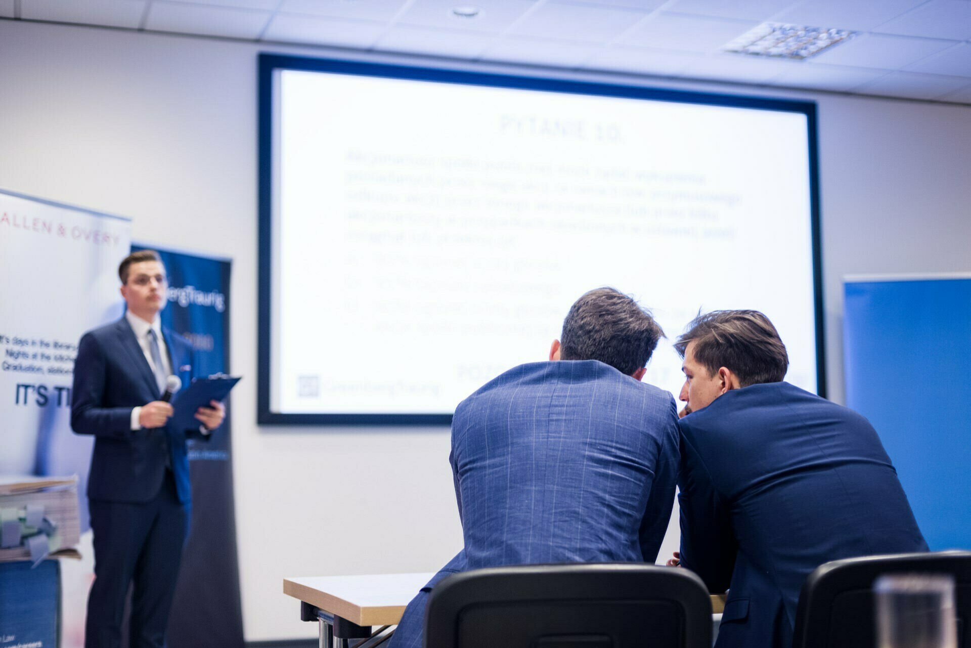 A presenter in a suit stands in front of a screen displaying a presentation and addresses the audience. In the foreground, two men in suits sit at a table, leaning over to discuss something privately. The scenery is reminiscent of a professional conference room, ideal for photo coverage of events.  