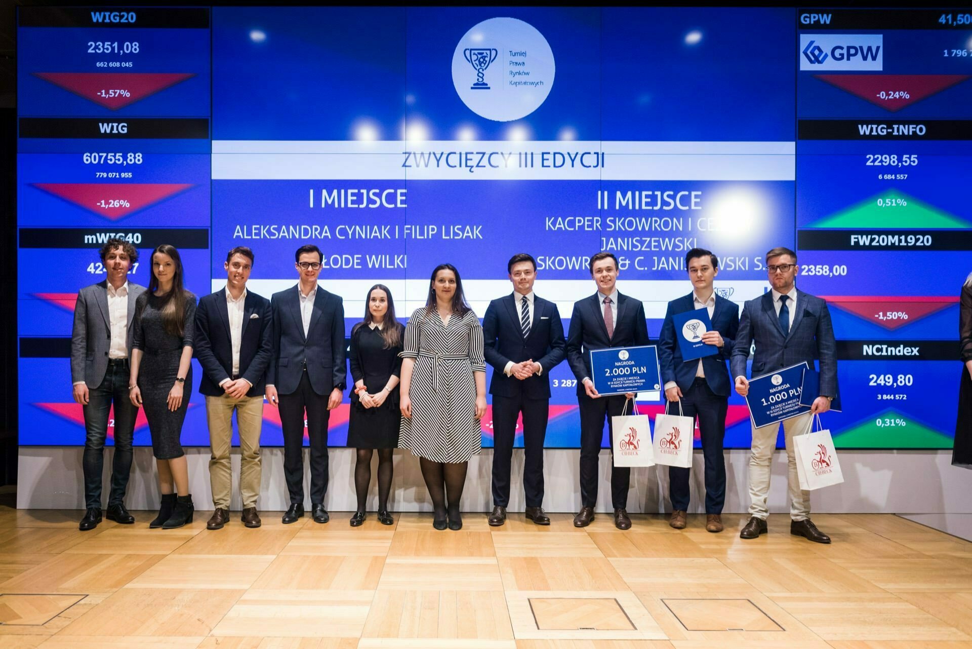 A group of people stand on a stage in front of a large screen displaying various information. Several people hold certificates and prize envelopes. A total of nine adults dressed in formal attire pose for an event photo.  