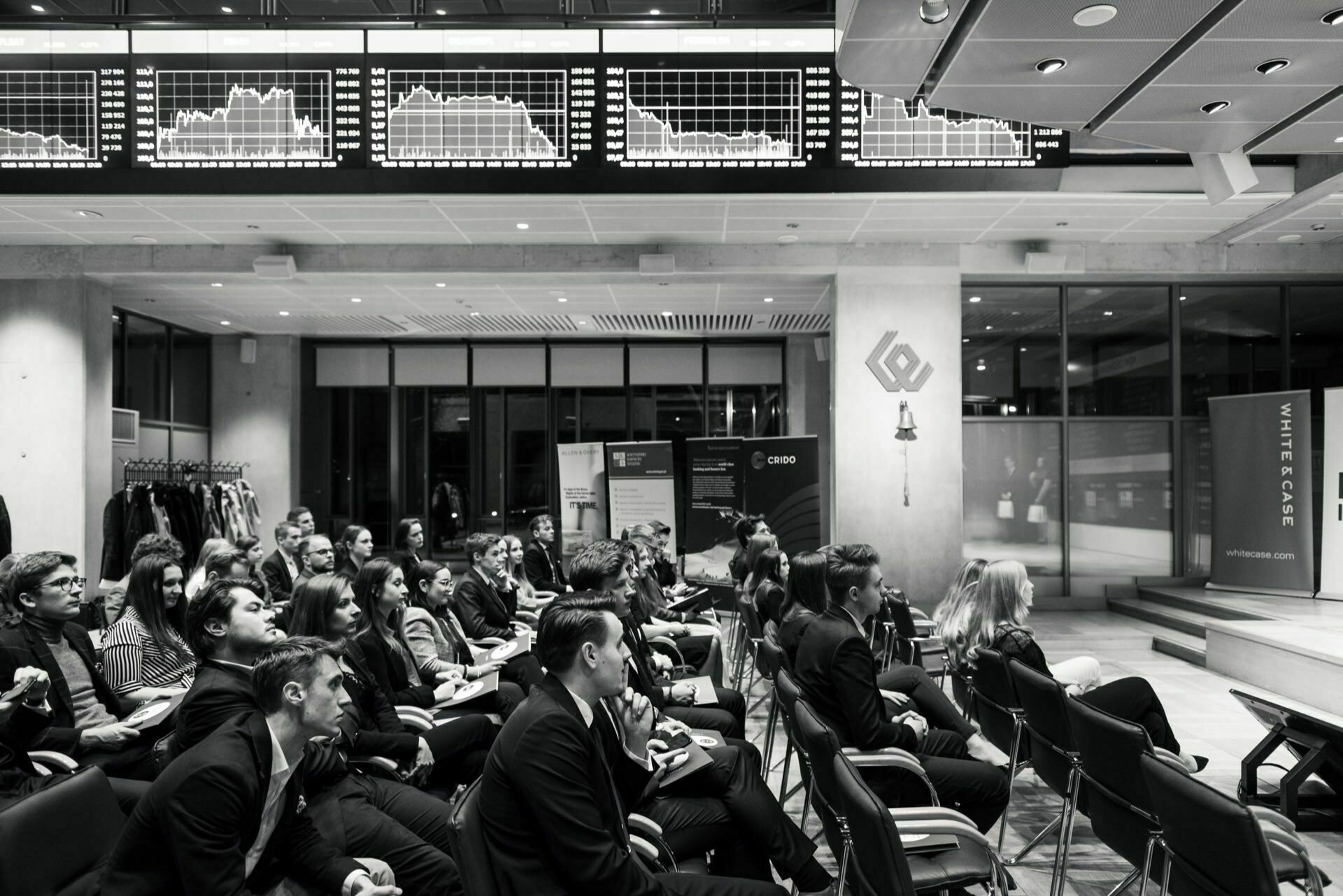 A black and white photo of an audience sitting in rows facing a screen in a modern room. Above them, numerous stock market charts are displayed on large monitors. Business attire dominates, and the atmosphere suggests a setting for a celebration or corporate event, perfect for an event photo shoot by event photographer warsaw.  