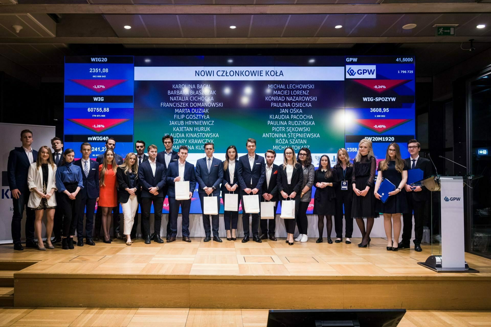 On the stage, a group of people dressed in formal attire stand in line, holding documents and awards. Behind them is a large digital screen displaying various names and numbers. The setting appears to be a formal event or ceremony, ideal for photo coverage of events.  