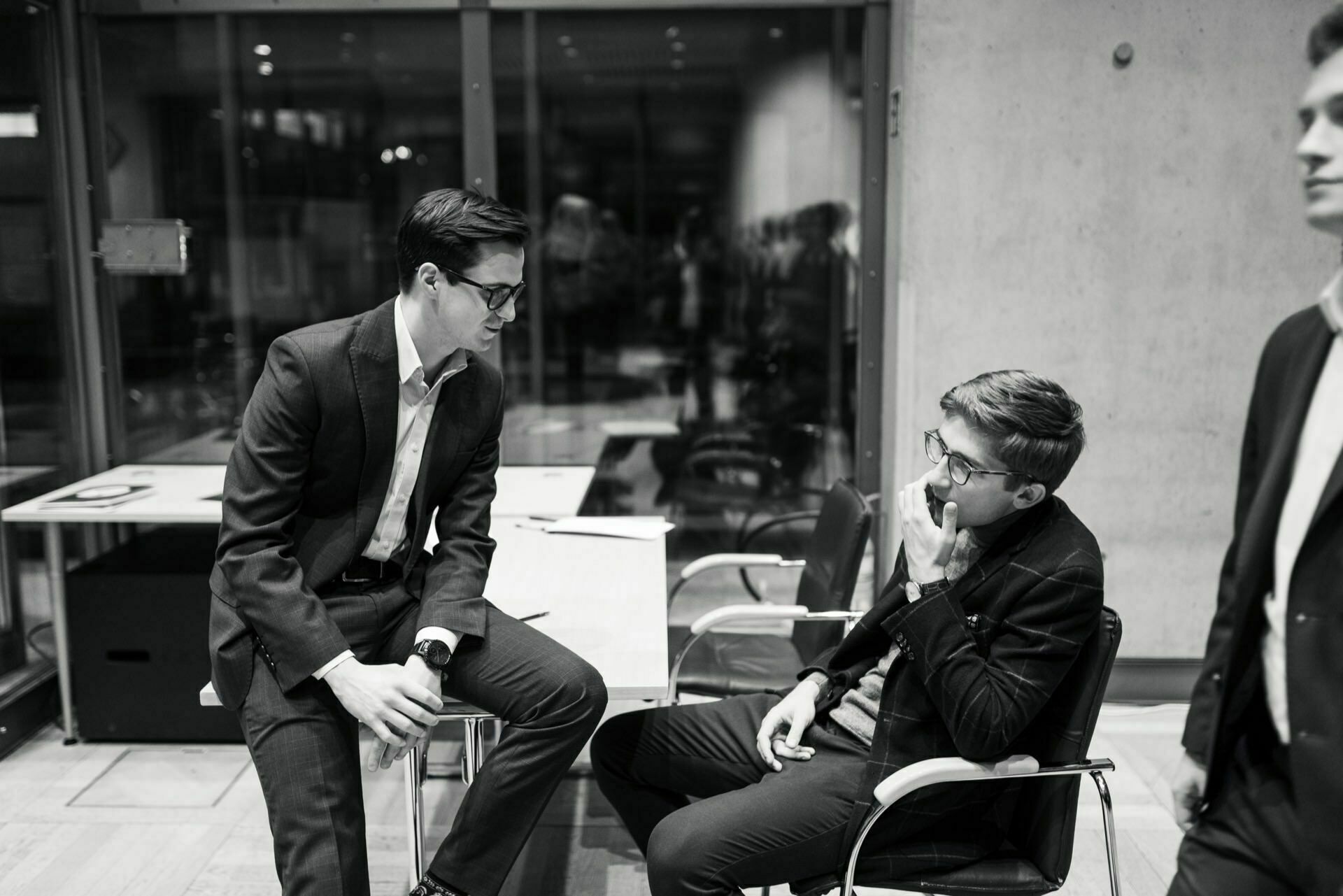 The black-and-white image shows three men in suits talking. Two are seated at a table; one is gesticulating and the other is listening intently. The third man stands slightly blurred at the edge of the frame. The setting appears to be a modern office, captured as part of a photo essay of an event.   