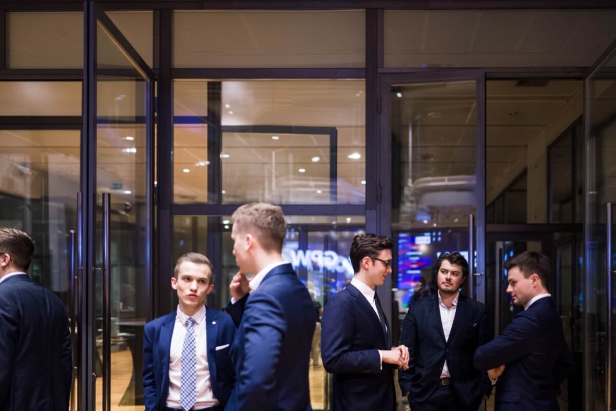 Eine Gruppe von fünf Männern in Anzügen unterhält sich in einem Gebäude, das an ein Bürogebäude oder ein Konferenzzentrum mit Glaswänden und Türen erinnert. Einige sind mit dem Gespräch beschäftigt, andere schauen sich um oder starren auf ihre Telefone - perfekte Momente für die Eventfotografie von Eventfotograf Warschau.