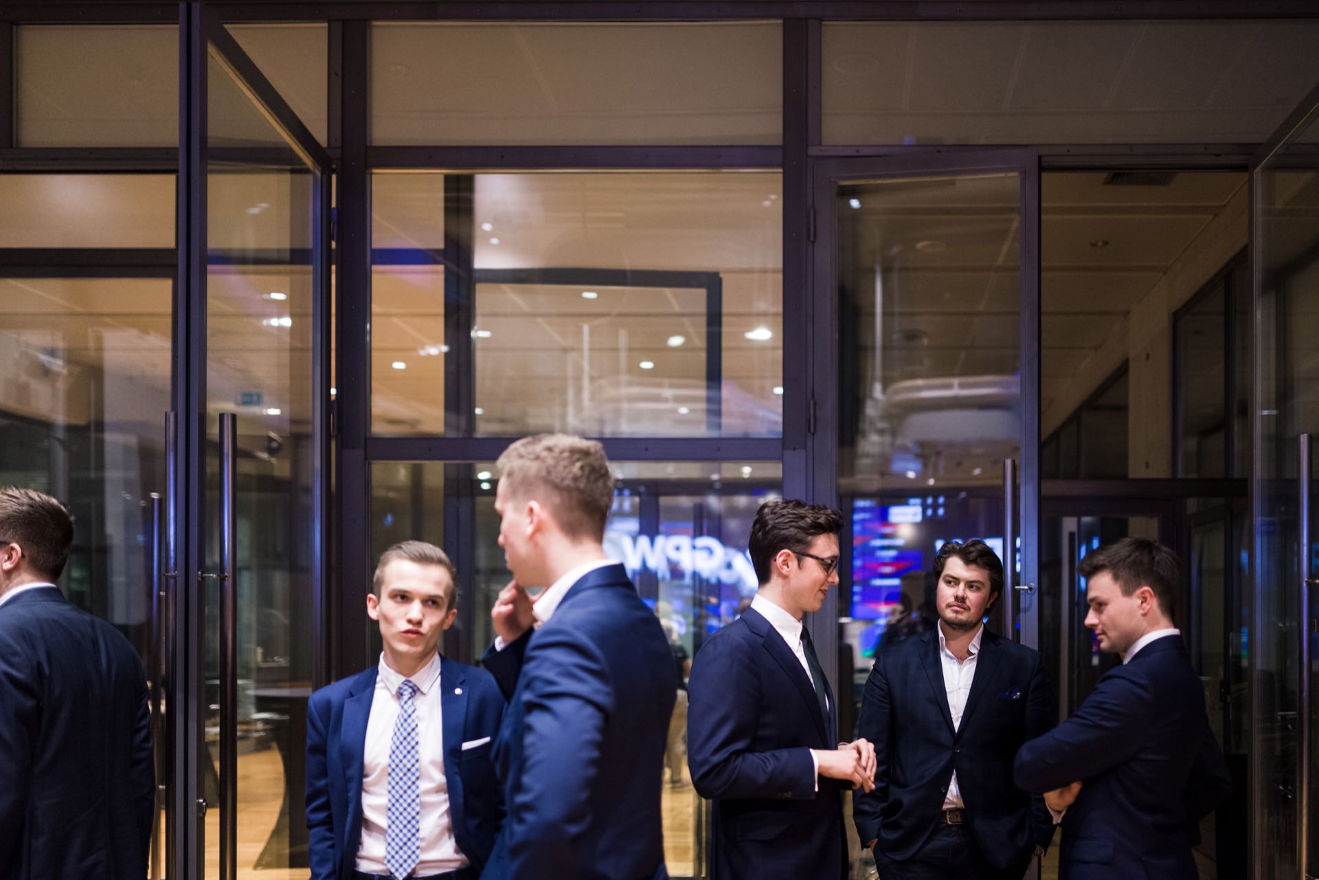 A group of five men dressed in suits are having a conversation inside something that resembles an office building or conference center with glass walls and doors. Some are busy talking, others are looking around or staring at their phones - perfect moments for event photo coverage by event photographer Warsaw. 