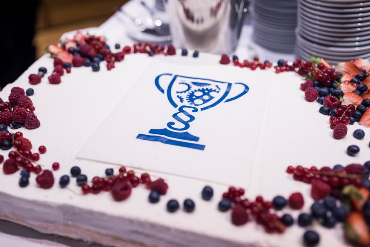 Ein rechteckiger Kuchen, der mit weißem Zuckerguss verziert und von verschiedenen Beeren umgeben ist. Die Torte zeigt eine Trophäe mit Ritzeln und einer DNA-Helix obenauf, gedruckt in Blau auf weißem Hintergrund. Im Hintergrund sind Teller und Besteck zu sehen, ideal für Event-Fotoshootings.