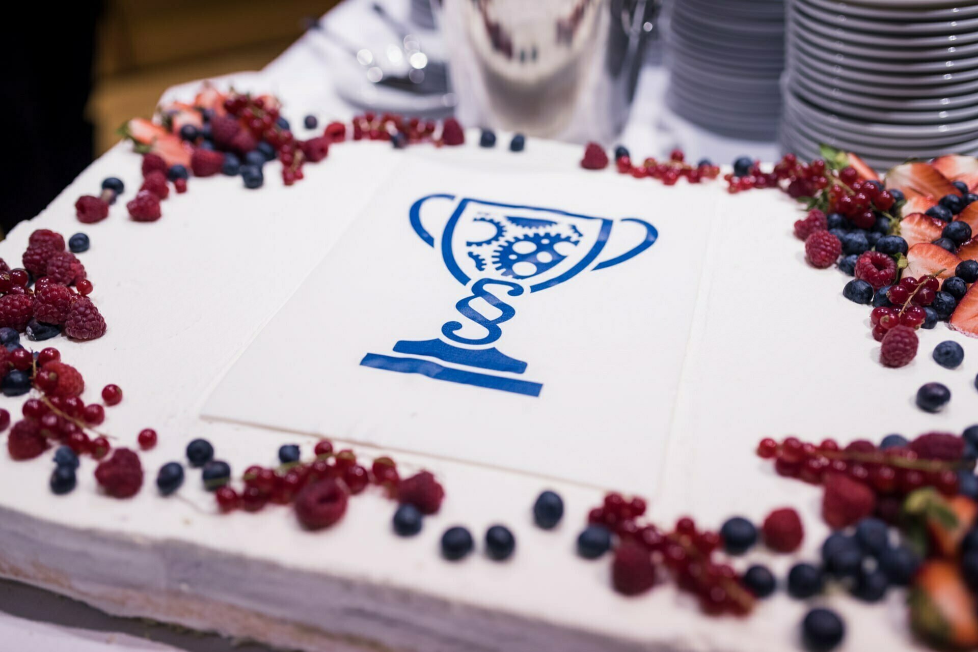 Rectangular cake decorated with white icing and surrounded by various berries. The cake depicts a trophy with gears and a DNA helix on top, printed in blue on a white background. Plates and cutlery are visible in the background, perfect for event photos.  