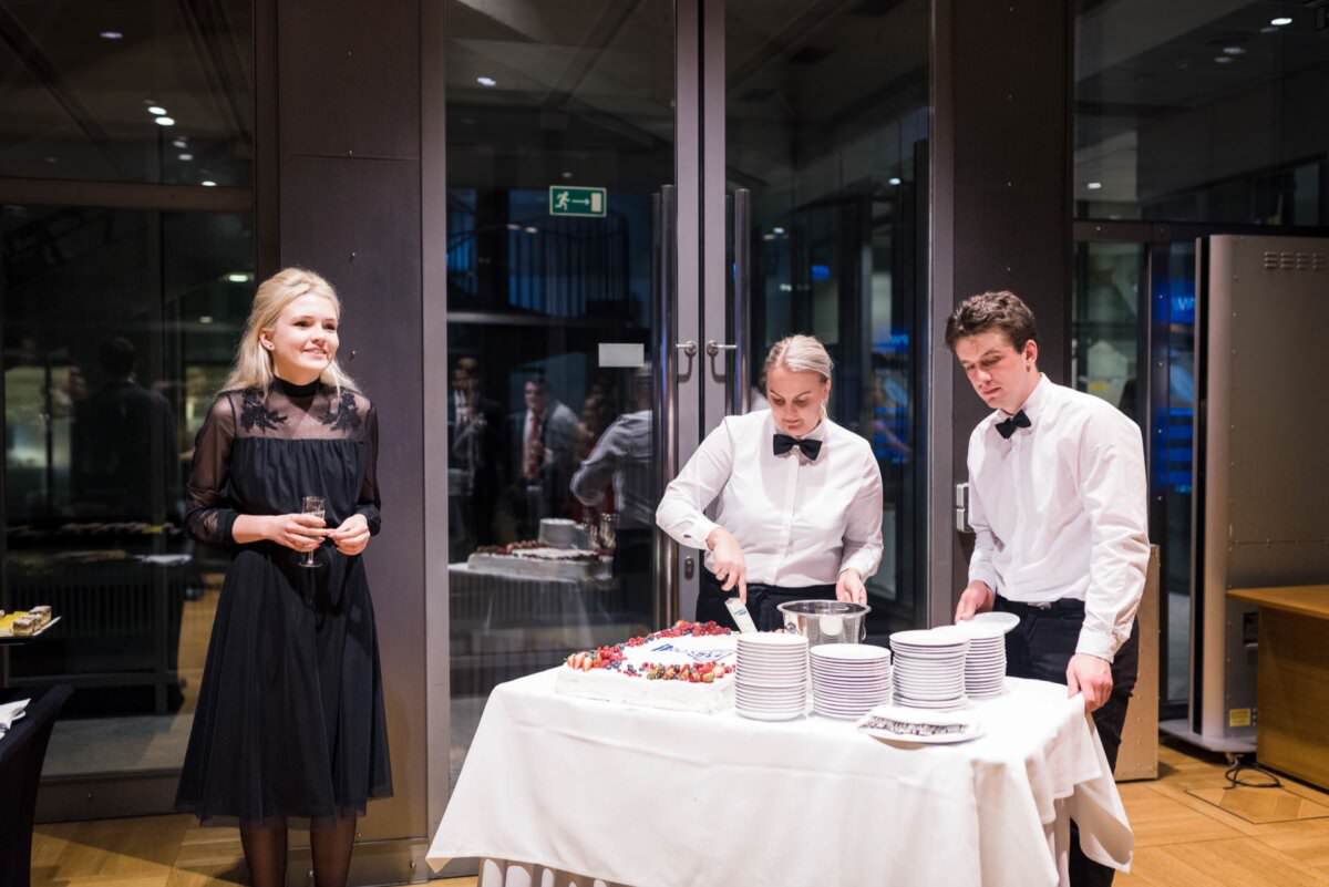 Links steht eine lächelnde Frau in einem schwarzen Kleid, während zwei Kellnerinnen in weißen Hemden und schwarzen Fliegen eine Torte auf einem mit einem weißen Tischtuch bedeckten Tisch anschneiden. Teller und Besteck sind ordentlich neben ihnen angeordnet. Im Hintergrund befindet sich eine große Glastür, die den perfekten Moment für die Eventfotografie einfängt.