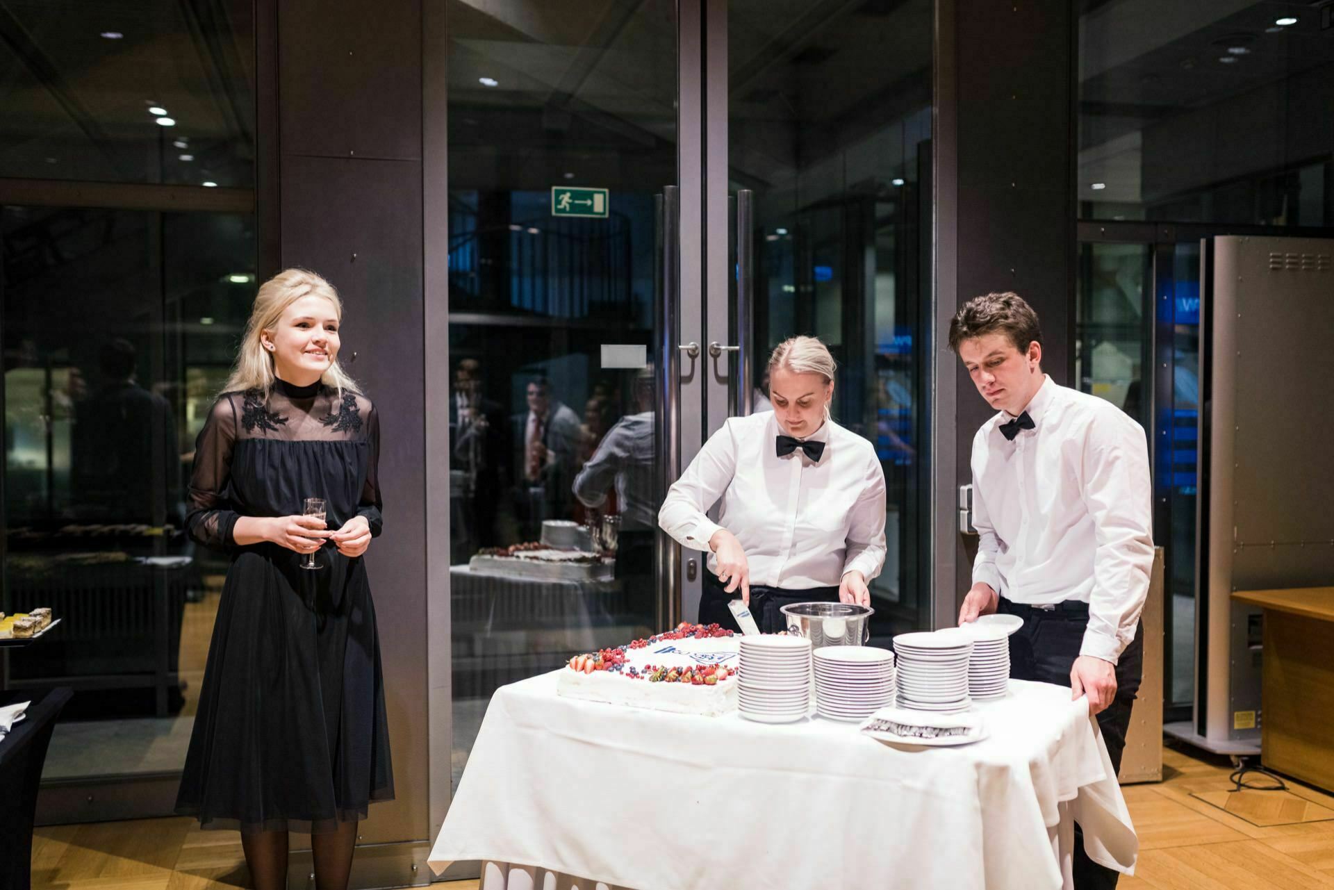 To the left stands a smiling woman in a black dress, while two waitresses in white shirts and black bow ties cut a cake on a table covered with a white tablecloth. Plates and cutlery are neatly arranged beside them. In the background is a large glass door that captures the perfect moment for event photography.  